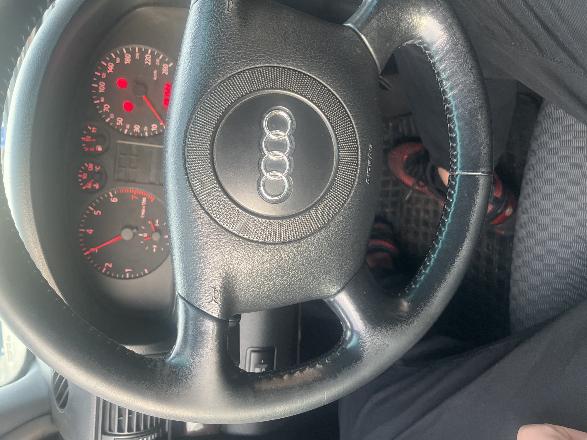The image shows the steering wheel of an Audi car, specifically the older model with the four-ring logo on the airbag cover. The dashboard is partially visible, showing the speedometer at around 20 km/h and the RPM gauge at around 3000. The driver's legs are visible, wearing dark pants and red-and-black athletic shoes. The overall environment suggests the car is in motion, as indicated by the speedometer and the angle of the shot. There is no information to infer about the location in the city of Sofia, Bulgaria.