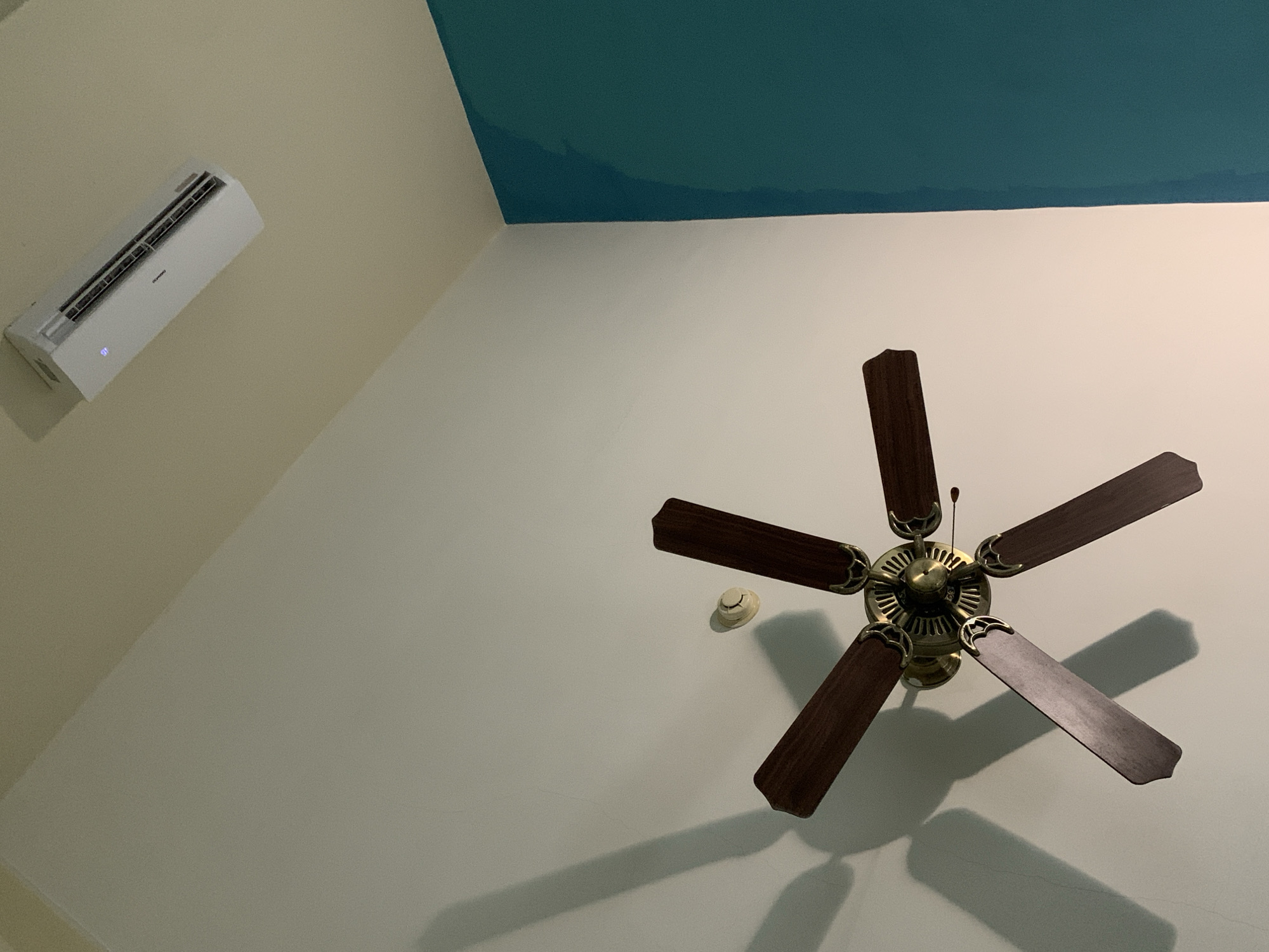 The image presents an upward-looking interior view of a room located in Playa El Agua, Venezuela. The ceiling is white, from which a brass-finished ceiling fan with five dark wooden blades is centrally suspended, casting distinct shadows. A white, circular smoke detector is affixed to the ceiling to the upper right of the fan's hub. The lower section of a wall on the left side of the frame is painted a vibrant teal-turquoise. The upper section of an adjacent wall on the right side of the frame is painted a light beige/cream, where a white split-system air conditioning unit is mounted high. The air conditioning unit exhibits a faint blue illumination from its digital display. The scene is illuminated by an artificial light source, creating sharp shadows.