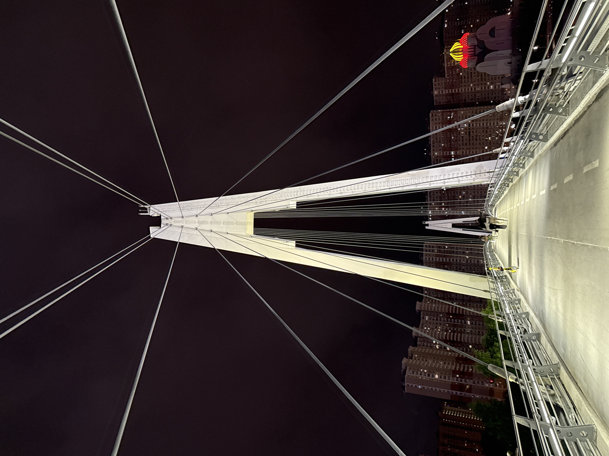 This is a night-time, wide-angle, low-angle, full shot of the pedestrian cable-stayed bridge in Khimki, Russia, located over the MKAD (Moscow Ring Road). The bridge is illuminated, with bright white lights highlighting its structure and pathways. A tall, white, central pylon dominates the center of the frame, from which numerous thin, taut cables extend outwards and downwards, creating a symmetrical, star-like pattern against the dark night sky.
The bridge deck is a wide, concrete walkway with visible lane markings and illuminated railings. In the distance, on either side of the bridge, are tall, multi-story apartment buildings, their windows lit, suggesting a densely populated urban environment. To the right of the frame, a domed building, possibly a church, is prominently lit with red and yellow lights.
On the bridge itself, two couples are walking away from the camera. One couple is closer to the foreground and appears to be walking in the same direction. The second couple is further down the bridge and to the left. The scene is clean and modern, with no visible debris. The overall impression is one of urban nighttime beauty and engineering.