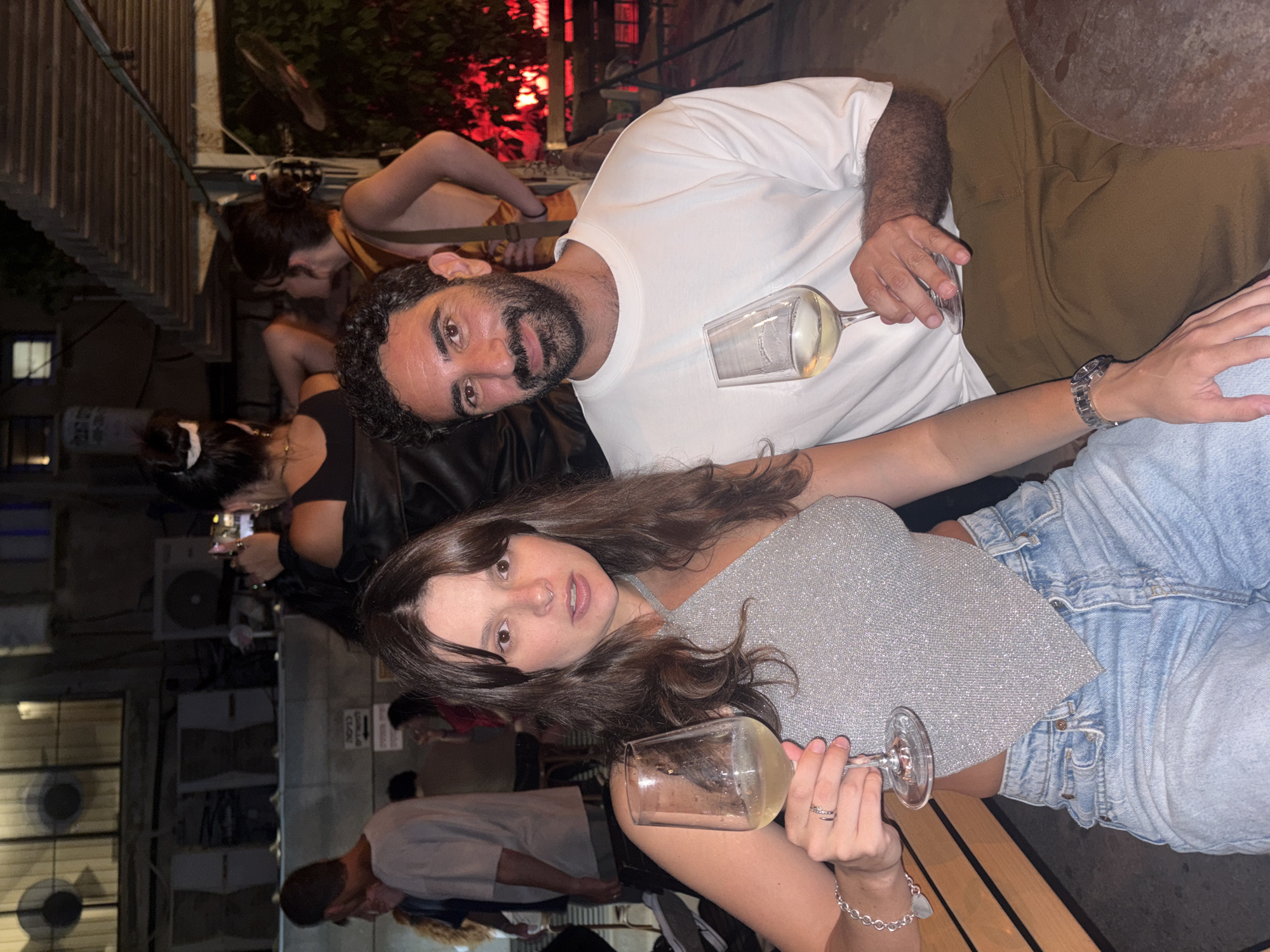 A photograph taken at night, likely in an outdoor urban setting in Tel Aviv, Israel, shows two people sitting at a table, holding wine glasses. The woman in the foreground, to the left, has long brown hair and is wearing a silver, sparkly halter top and blue jeans. She is holding a wine glass in her left hand and appears to be looking at the camera. The man beside her, to the right, has dark curly hair and a beard, wearing a white t-shirt and brown pants. He is smiling and also holding a wine glass. In the background, other people and a dimly lit urban scene with some vegetation are visible.