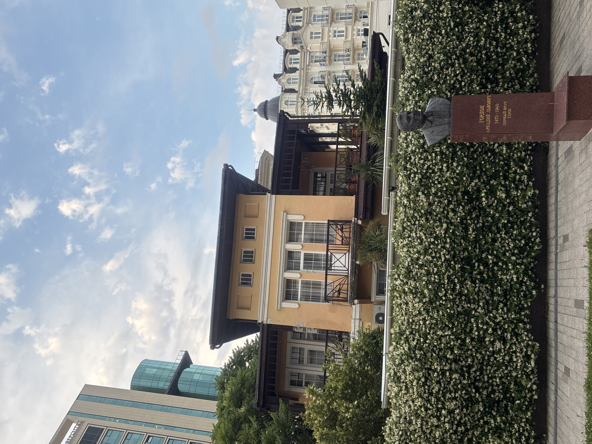 A bust of Arkady Lvovich Gordon, a physician from 1871-1940, stands on a stone pedestal in front of a dense, blooming hedge. The bust is located in front of a light yellow building with a dark roof and balconies. Behind the yellow building, a modern glass skyscraper and a white building with ornate architecture are visible. The sky is partly cloudy. The scene is located in Sochi, Russia.