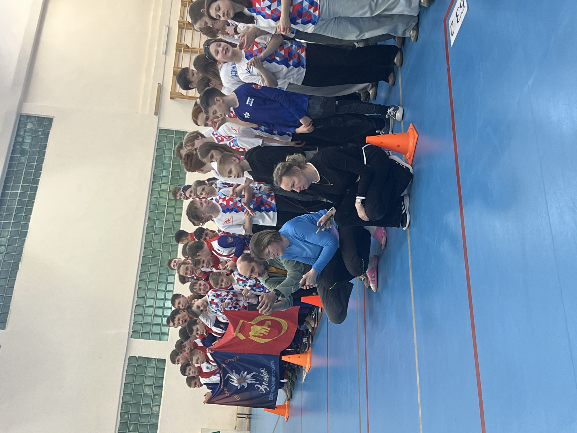 A long line of children and adults stand on a blue, indoor track, presumably in Berezniki, Russia, based on the writing on the flags. The floor has orange lines marking lanes, and orange cones are scattered along the edge. The group is oriented horizontally across the image's bottom edge, facing the viewer from left to right. Many children wear matching red, white, and blue checkered shirts or blue shirts. Two individuals, dressed in black and light blue, are crouched down, seemingly interacting with the group, possibly taking photos or giving instructions. Two flags are held by the group in the front; one is red with a fist emblem, and the other is darker with a flower and writing. The background shows a gymnasium or sports hall with high ceilings and windows. The lighting is bright.