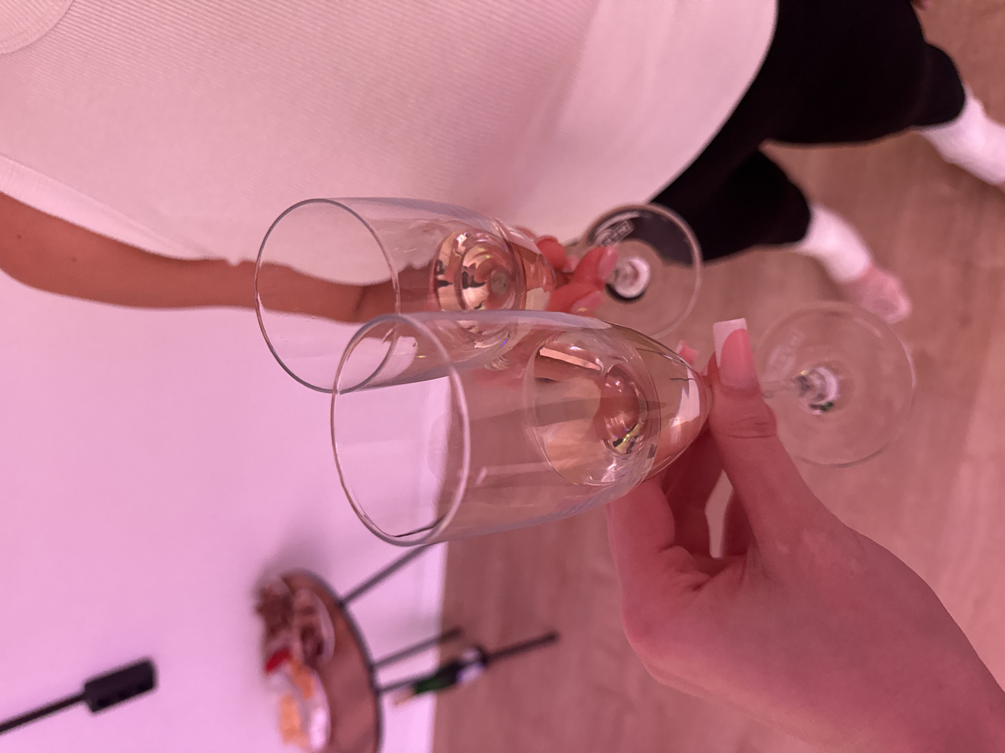 In the foreground of this image, a person's left hand, with manicured pink nails, holds two champagne flutes. The flutes contain a light-colored liquid, presumably champagne or sparkling wine. The flutes are positioned in a toasting gesture. In the background, out of focus, another person's torso is visible, wearing a white ribbed top. The environment appears to be indoors, with a pinkish hue cast over the scene, suggesting artificial lighting. A small table with snacks is visible to the left, and the floor is wooden. The scene is set in Zakharovo, Russia.