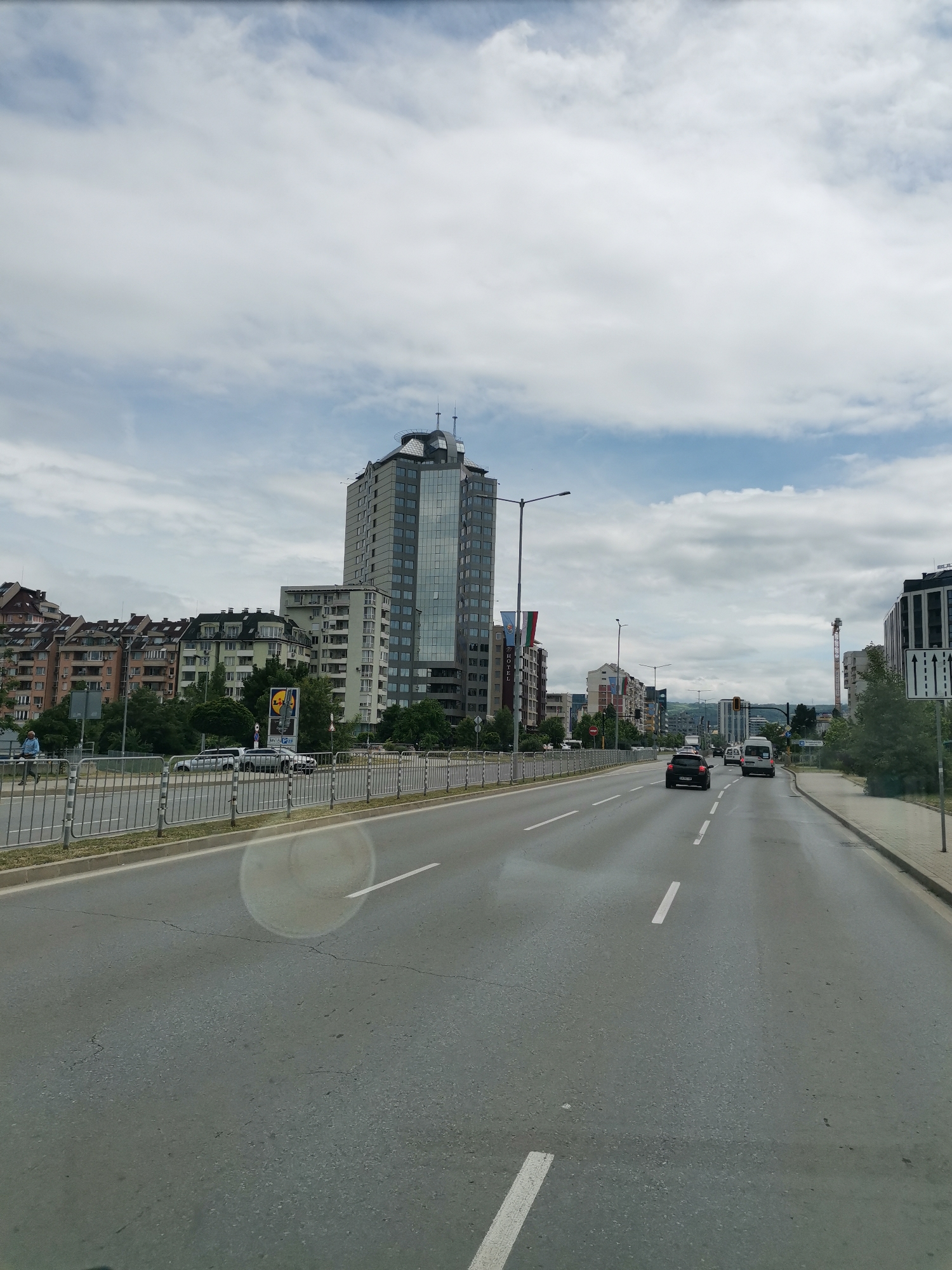 The image depicts a wide, multi-lane asphalt road with white lane markings, viewed from a vehicle. The road appears to be in an urban environment, likely in Sofia, Bulgaria, given the presence of distinct architectural styles and Bulgarian flags visible on a building.
On the left side of the road, a metal guardrail separates the traffic lanes from a sidewalk where a lone individual in a light blue shirt is walking. Beyond the sidewalk, a line of multi-story apartment buildings with various architectural features, including angled roofs and balconies, stretches into the distance. A prominent, modern, glass-fronted skyscraper stands out among these buildings, topped with antennas. A large billboard for "Lidl" is also visible near the apartment buildings.
On the right side of the road, more modern buildings and trees line the street. Traffic is flowing, with several cars and a white van visible on the road ahead. The sky is overcast with large, white clouds.
The overall scene captures a typical urban streetscape in Sofia, characterized by a mix of residential and commercial buildings, modern infrastructure, and active city life. The perspective suggests the photo was taken from a moving vehicle, looking forward along the road.