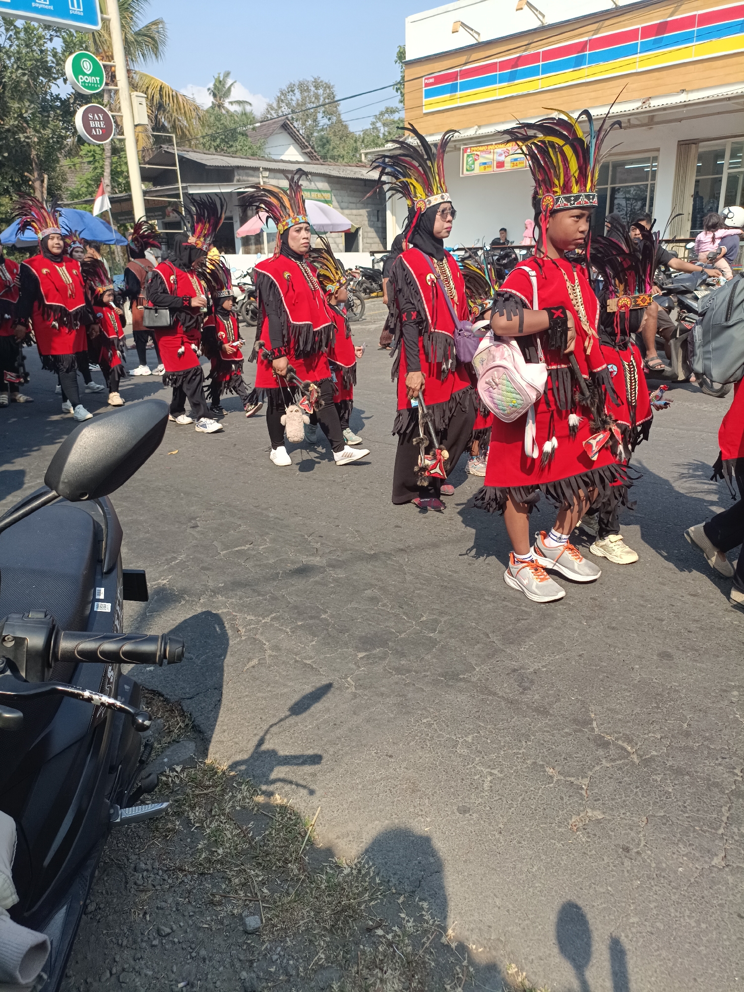 In Jatitengah, Indonesia, a group of individuals, predominantly children and young adults, are participating in a parade or cultural event. They are adorned in vibrant red and black costumes featuring elaborate feathered headdresses. The costumes are decorated with fringe and adornments that suggest traditional or ceremonial attire. The participants are walking on an asphalt road, with a motorcycle visible in the foreground and a building with a colorful striped facade in the background, identifying the location as a commercial area. The weather appears to be sunny, casting shadows on the road. The scene captures a moment of cultural celebration within the urban landscape.