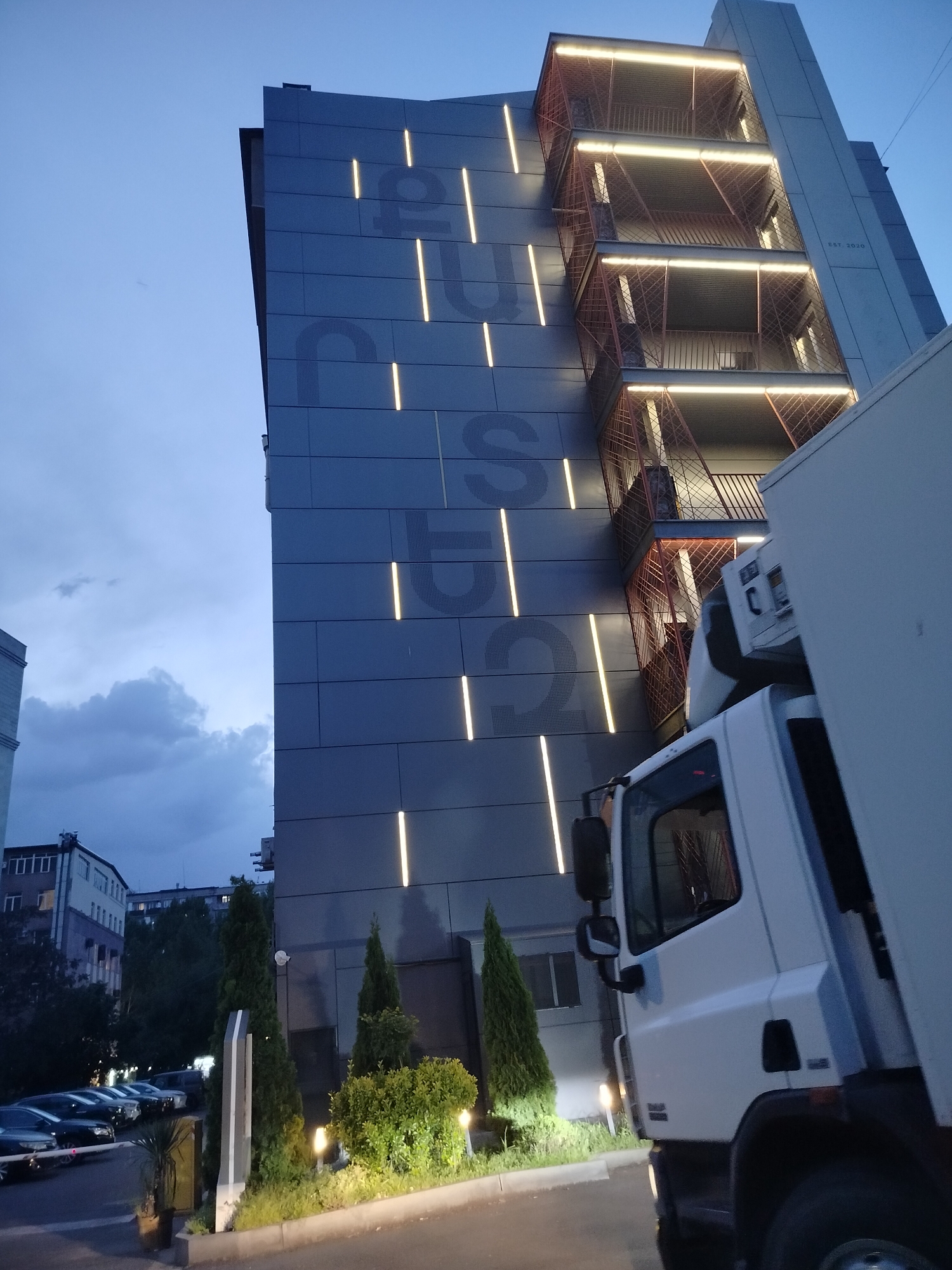 The image shows a view of a modern building in Yerevan, Armenia, at dusk. The building is characterized by its grey facade, embellished with vertical light strips. The word "EUST" is subtly embedded in the facade design. An external metal staircase with illuminated landings is on the right side of the building. A white delivery truck is parked near the building, partially obstructing the view. In the background, a cityscape with residential buildings and overcast sky is visible. The foreground features neatly trimmed greenery with decorative lighting. There is a parking area to the left with parked cars behind a gate.
