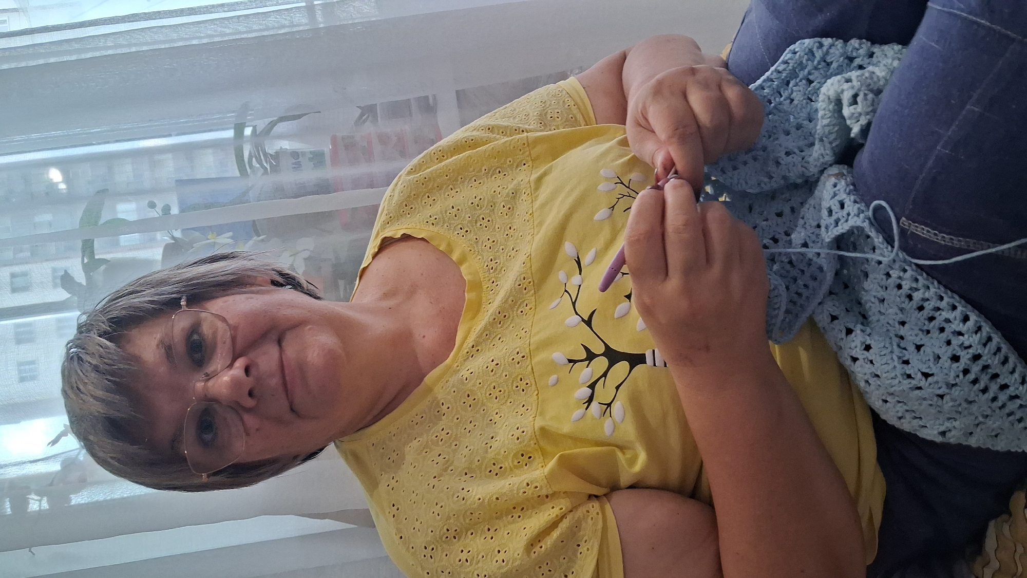 A medium shot shows a woman with short, graying hair and glasses, wearing a yellow t-shirt with a white tree design. She is sitting and crocheting a light blue fabric with a purple crochet hook. She is looking towards the camera with a slight smile. The background consists of a sheer white curtain, a window, and what appears to be apartment buildings visible through the curtain. The lighting is soft, suggesting an indoor environment. The image was taken at an undisclosed location within the city of Koltsovo, Russia.