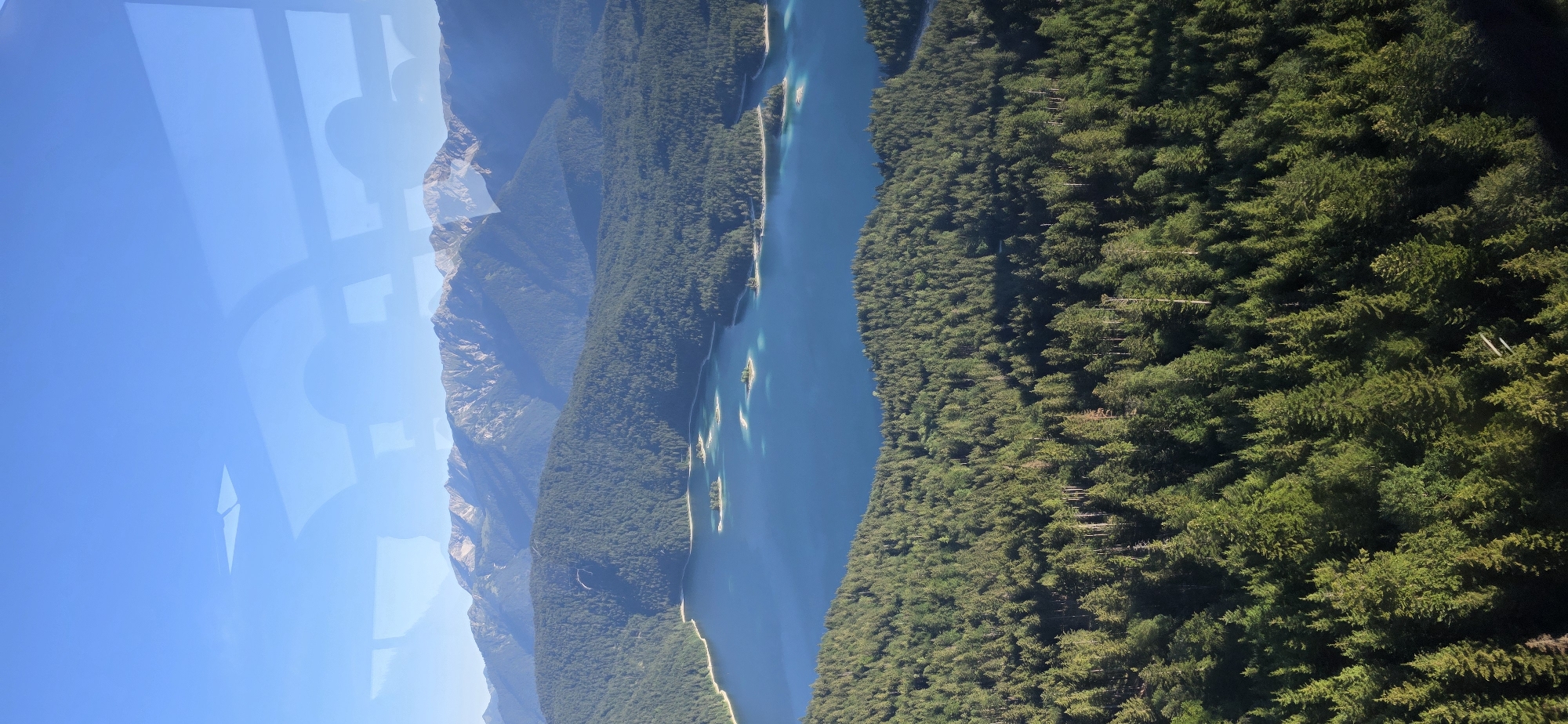 This is an aerial photograph of the Eibsee lake in Grainau, Germany, on a clear day. The lake is a vibrant blue and dotted with several small islands. It is surrounded by a dense, dark green forest that extends up the slopes of the mountains in the background. The mountains are rugged and rocky, with patches of snow visible on their peaks. The sky is a brilliant blue with some faint reflections of what appear to be windows or a skylight. The image captures a paraglider in the air above the forest and lake.