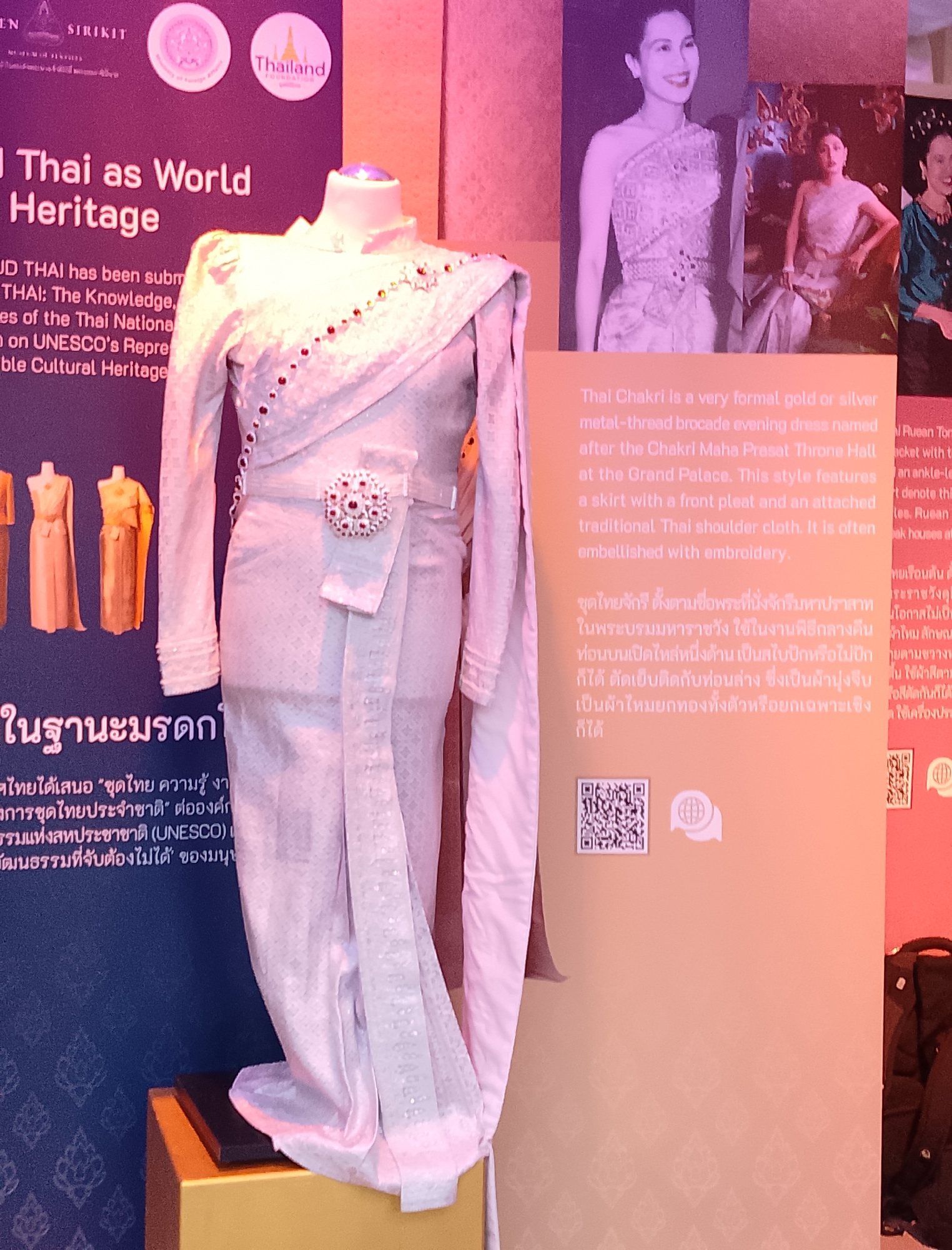 An exhibit displays traditional Thai clothing, prominently featuring a pale pink, intricately patterned Chakri outfit on a mannequin. The outfit includes a long-sleeved top with a high collar and a wrap-around skirt adorned with a decorative brooch. Beside the mannequin, a large poster displays text in both English and Thai, describing "Thai as World Heritage" and the Chakri dress. Smaller images of women in similar attire are also visible on the poster. To the right, a smaller display showcases additional Thai garments. The overall environment appears to be an indoor exhibition space with soft, ambient lighting.
