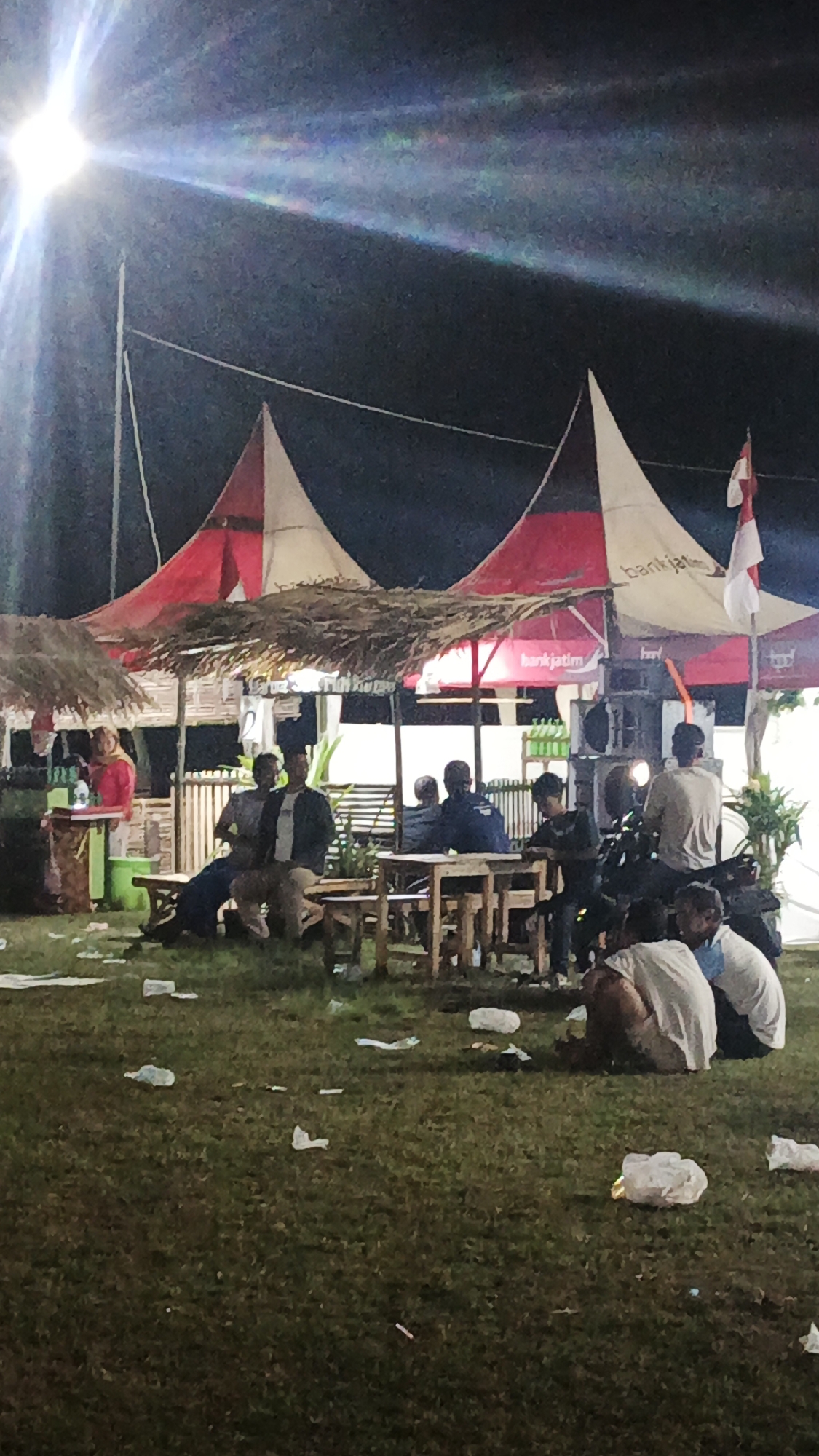 In the city of Ketemasdungus, Indonesia, a nighttime outdoor gathering is underway. Several individuals are seated at tables and on the ground, suggesting a casual social event. The scene is illuminated by a bright, artificial light source, possibly a spotlight or stage light, which creates lens flares in the upper left quadrant of the image. In the background, there are tents with red and white triangular designs, and one tent has "bankjatim" written on it, indicating a potential sponsorship or presence of Bank Jatim. An Indonesian flag is visible on a pole attached to one of the tents. In the foreground and scattered across the grassy area are various pieces of litter, including plastic bags and wrappers, suggesting the event has been ongoing and is not yet cleaned up. The overall atmosphere appears relaxed, with people engaged in conversation or simply resting outdoors.