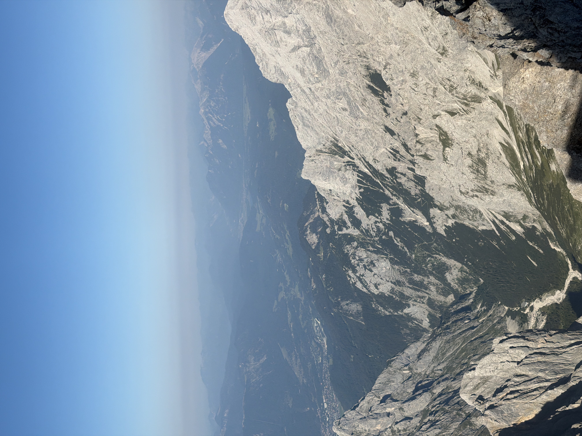 The image is an aerial view of a mountainous region, likely taken from a high vantage point. In the foreground, a rugged, rocky mountain face dominates the right side of the frame. Its surface is textured with numerous cracks and crevices, and it casts a deep shadow to the lower left. To the left of this prominent rock formation, a steep, dark valley descends, filled with dense green coniferous forest. A dry, rocky riverbed snakes its way through the valley floor.
Further into the distance, the landscape unfolds into a series of rolling mountain ranges, becoming progressively hazier and bluer with atmospheric perspective. A small town or village with buildings and roads is visible nestled in a valley between the closer mountains and the distant ranges. The sky above is a clear, bright blue, with a faint haze near the horizon.
Given the context of Garmisch-Partenkirchen, Germany, this view is likely of the Zugspitze or a neighboring peak, looking out over the surrounding Bavarian Alps. The town visible in the mid-ground could be Garmisch-Partenkirchen itself or a smaller settlement within the valley. There are no people or specific actions depicted in the image. The environment is natural, characterized by mountains, forests, and a clear sky.