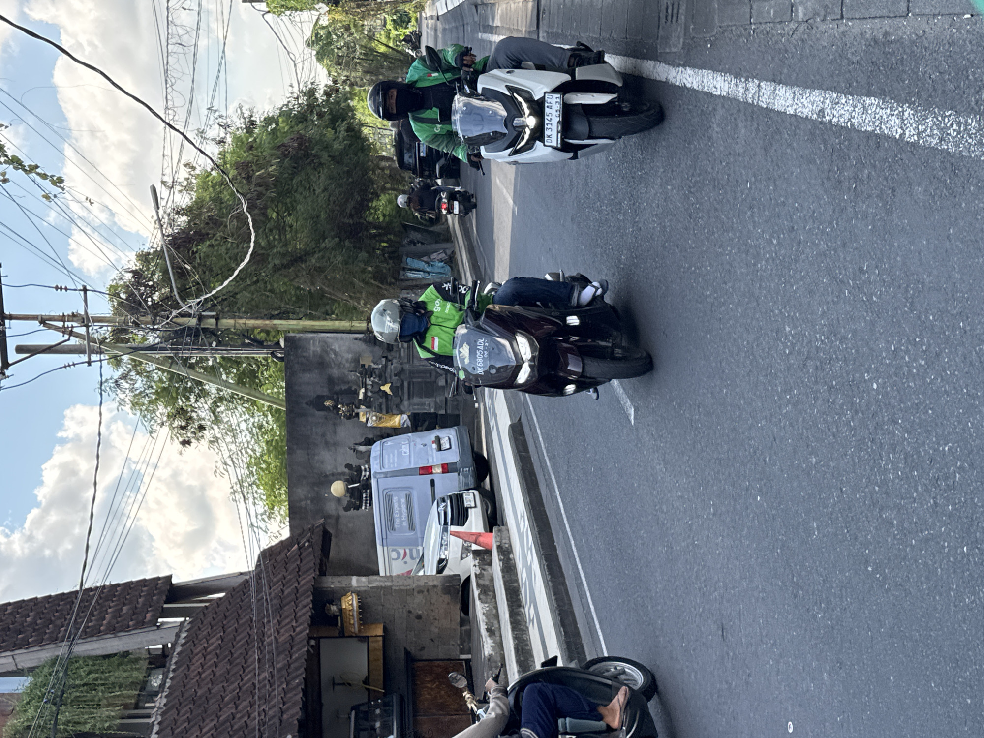 This daytime street scene captures an active road in what appears to be an urban or semi-urban setting, likely in Kuta, Indonesia, given the context clues. The bright sunlight and scattered white clouds indicate good weather.
Several people are visible on motorbikes, which dominate the traffic. In the mid-ground, two individuals are prominent on their scooters, both wearing green jackets and helmets. One is on a white motorbike with the visible license plate "DK 3145 AFU" and the dates "05-31". The other is on a darker motorbike, with "DK 6805 ADL" and "06-2" on its license plate. The green jackets, one partially displaying "go" text, suggest they are ride-hailing drivers (e.g., Gojek). Other motorbikes and riders are also present, including one in the foreground, with the rider's legs and feet, clad in sandals, visible.
Along the side of the road, a white van is parked or driving, clearly branded with "calmic" and the slogan "The Experts in Hygiene." A white car is behind it, and an orange traffic cone stands nearby. Buildings with distinctive brown tiled roofs line the street, partially obscured by trees and lush greenery. A network of utility poles and numerous overhead wires crisscrosses the sky, adding to the urban landscape. The road surface is asphalt, marked with white solid and dashed lines, indicating lanes or shoulders.