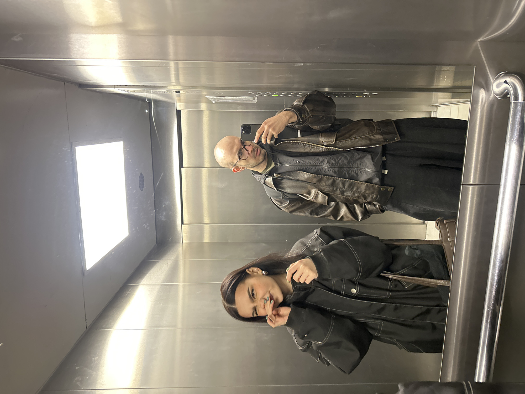 A medium, full shot captures two people in an elevator. The man in the reflection, located in the top half of the image, is bald, wearing glasses and a brown leather jacket over a dark shirt. He is holding a black smartphone, seemingly taking the photo. The woman in the reflection, in the bottom half of the image, has dark hair and wears a black jacket. She is smiling and holding her hand near her face. The elevator has stainless steel walls, a bright light fixture on the ceiling, and a metal handrail on the right side. The photograph appears to be a reflection in a mirror. Based on available information, the location is potentially Moscow, Russia.