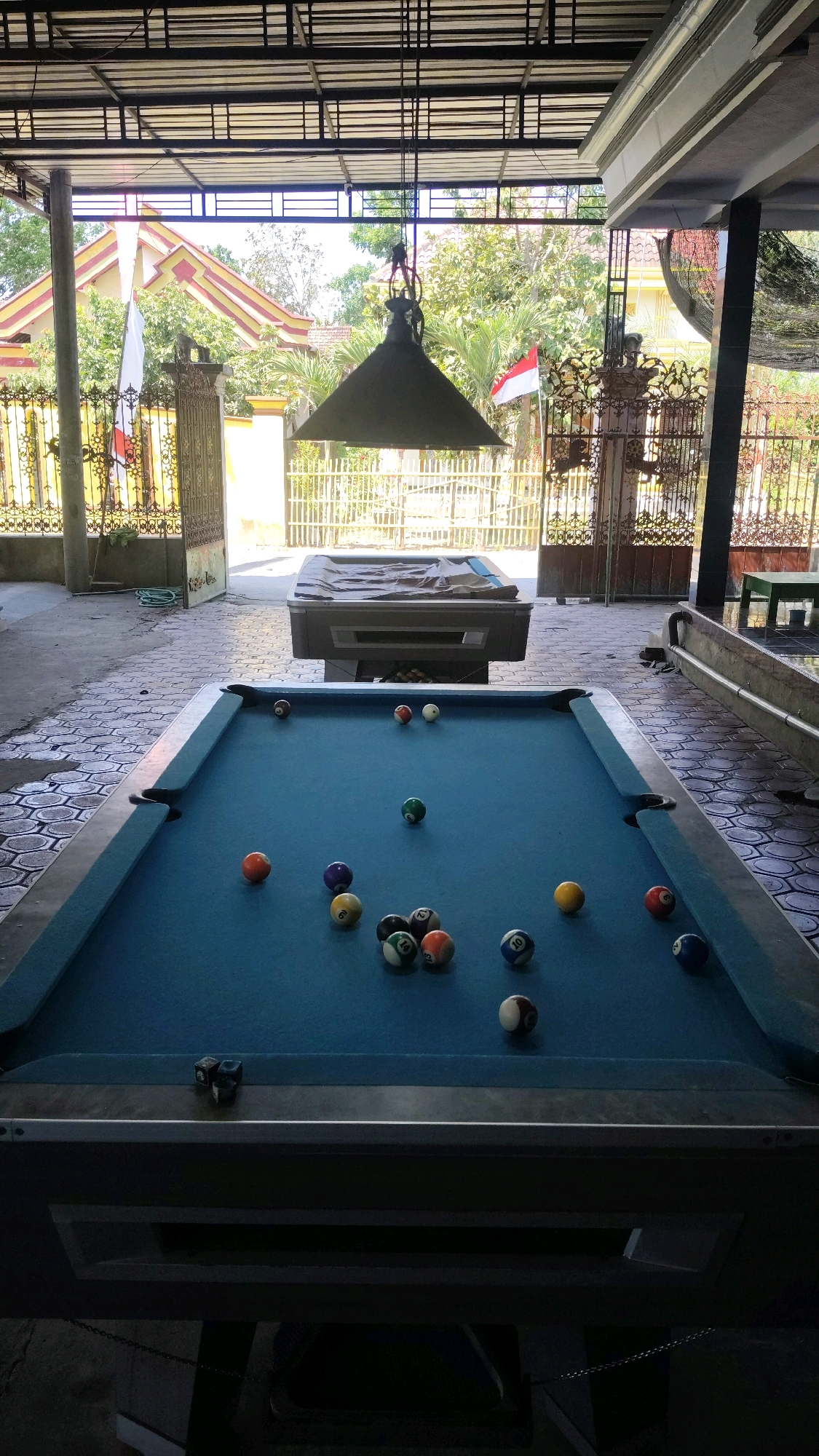 An outdoor billiard hall in Jabon, Indonesia features a blue felt pool table with several billiard balls scattered across it. The table is illuminated by a large, dark industrial pendant light hanging from the ceiling. In the background, a second, covered pool table is visible. The setting is open-air, with a corrugated metal roof supported by pillars. A tiled patio surrounds the pool tables, and beyond that, a fence encloses the area, revealing lush green trees and a house with a red and yellow roof. Two Indonesian flags are prominently displayed, one on a flagpole and another attached to a fence. The scene appears to be in a state of disuse, with some of the billiard balls seemingly out of place or not arranged for a game.