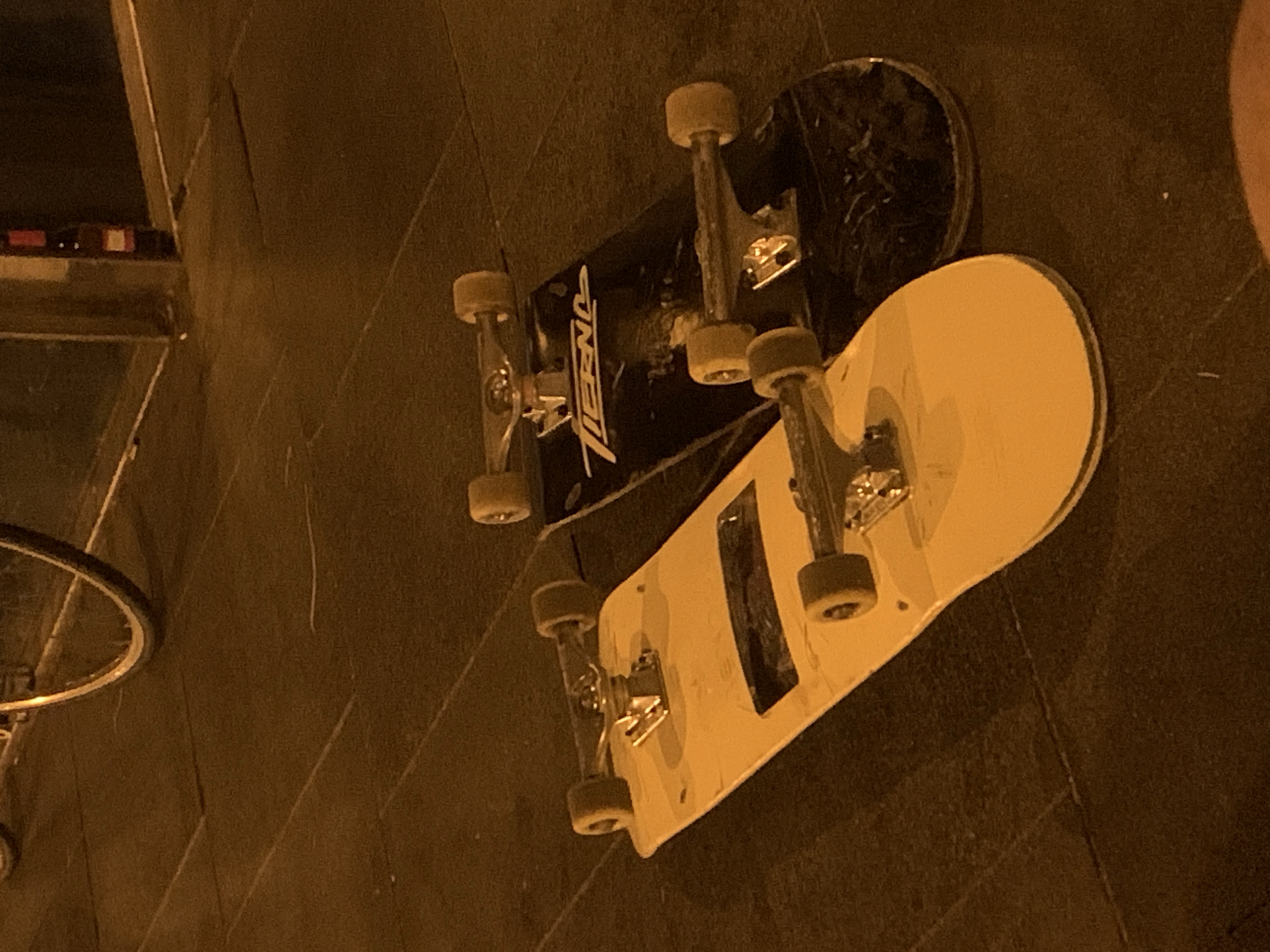 Two skateboards are positioned side-by-side on a dark, tiled ground surface, likely an outdoor urban setting during night or late evening due to the warm, dim lighting. The primary skateboard on the left is black with white text, clearly showing the brand "ONEILL" on its underside. It features metallic trucks and yellowish-beige wheels. Adjacent to it, on the right, is another skateboard, predominantly off-white or pale yellow, also equipped with metallic trucks and matching yellowish-beige wheels. This second skateboard has a darker, rectangular insert near its center.
In the upper left quadrant of the main image, and further detailed in a crop, a partial view of what appears to be a wall or structure is visible, with some blurry red and white rectangular objects, possibly lights or reflectors on a distant ledge. On the far left, a portion of a bicycle wheel and frame can be seen, partially resting against the ground. The ground itself is dark, showing faint patterns of tiles or paving slabs. No people are visible, and the skateboards appear to be at rest, suggesting a pause in activity or merely being stored. The overall mood is quiet and still, illuminated by the artificial light sources.