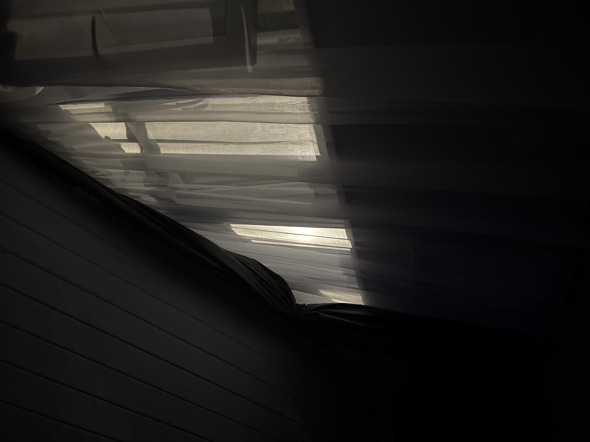 A dimly lit room features a window covered by sheer, white curtains. Sunlight streams through the window, creating bright vertical lines of light that are diffused by the fabric. To the left of the window, a dark, heavy curtain is partially drawn, obscuring the view and casting a shadow. The ceiling is visible in the upper left corner, appearing to be constructed of wooden planks. The overall impression is one of subdued natural light entering a private space. The location is described as Doma 1096 Km, Russia.