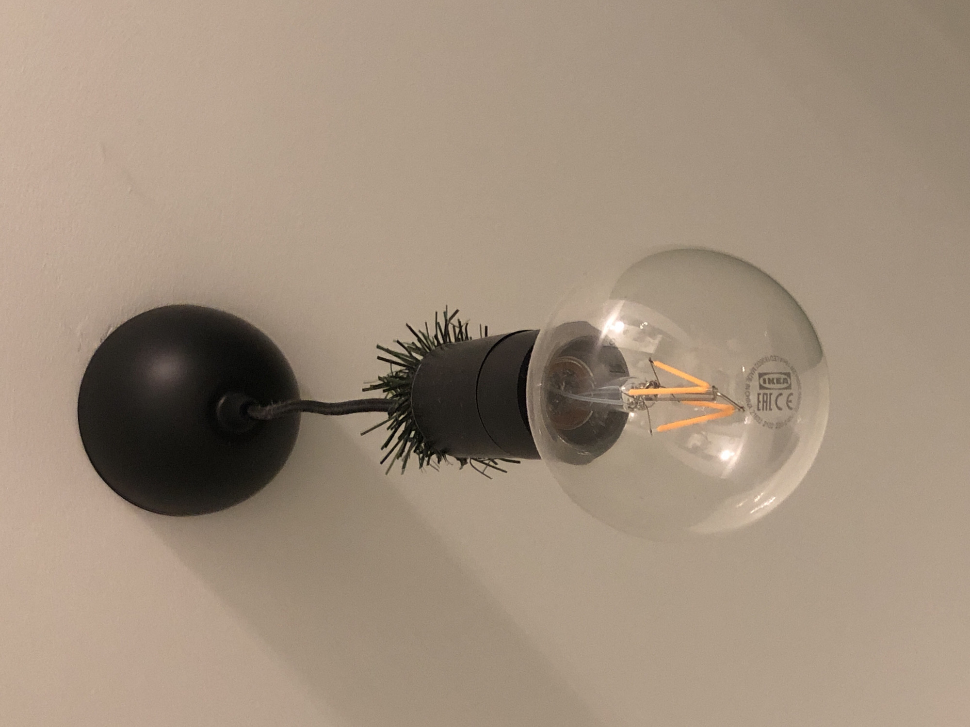 The image shows a close-up of a modern light fixture hanging from a ceiling. The fixture consists of a round, black canopy attached to the ceiling. A black cord extends from the canopy, leading to the light bulb assembly. The assembly is a black cylindrical holder with what appears to be artificial green foliage sticking out from the top. Below this, a clear, spherical light bulb is attached. Inside the bulb, an LED filament glows with a warm orange light. The bulb has some text and symbols printed on it, including "EAC" and "IKEA". The background is a plain, light-colored wall, possibly painted. There are no people or other identifiable objects or actions in the scene. The lighting suggests the photograph was taken indoors, likely in a residential or commercial setting.