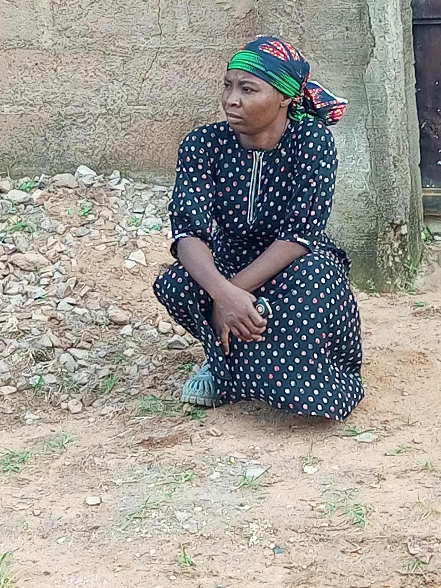 A woman is crouched on the ground outside. She is wearing a dark blue dress with white and pink polka dots, a colorful headscarf, and grey shoes. The woman is looking to her left and holding a small object in her right hand. The ground around her is dirt and small stones. Behind her is a textured wall and a tree trunk. The location is the city of Tempure, Nigeria.