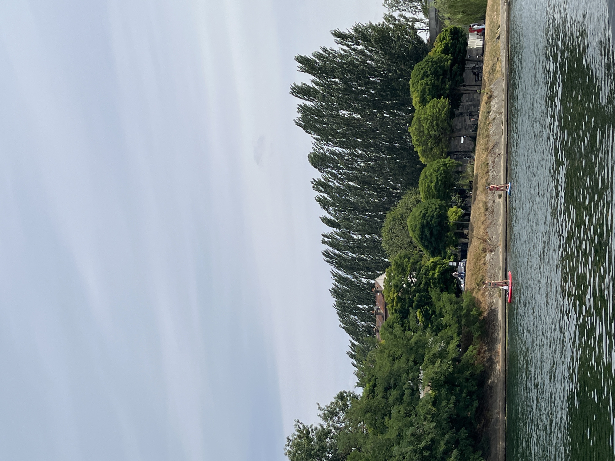 The image shows a wide river with two people paddleboarding. The riverbank is lined with trees, and in the background, there are buildings and more trees. The sky is overcast. This scene is located on the Marne River in Joinville-le-Pont, France, a town known for its water activities and guinguettes.