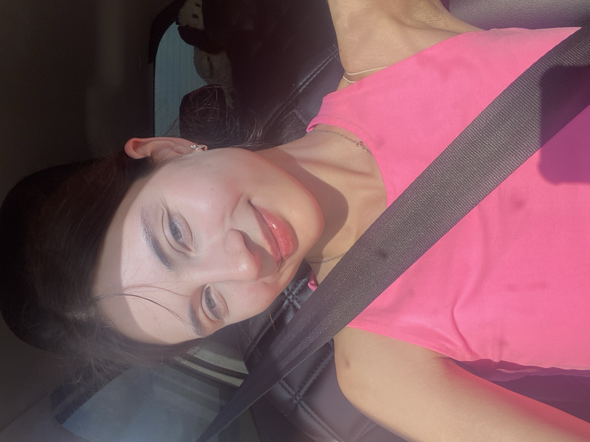 A young woman with dark hair and light blue eyes is captured in a selfie from inside a car. She is wearing a bright pink tank top and a silver necklace. A black seatbelt is prominently displayed across her chest. Sunlight streams in from the side, casting a warm glow on her face and highlighting her features. Her lips are slightly parted in a smile, and she appears to be looking directly at the camera. The background is blurred, but suggests the interior of a vehicle with dark upholstery and a window. The image is taken during daylight hours. There is no information to determine the location in the city of Kamkoye, Russia.