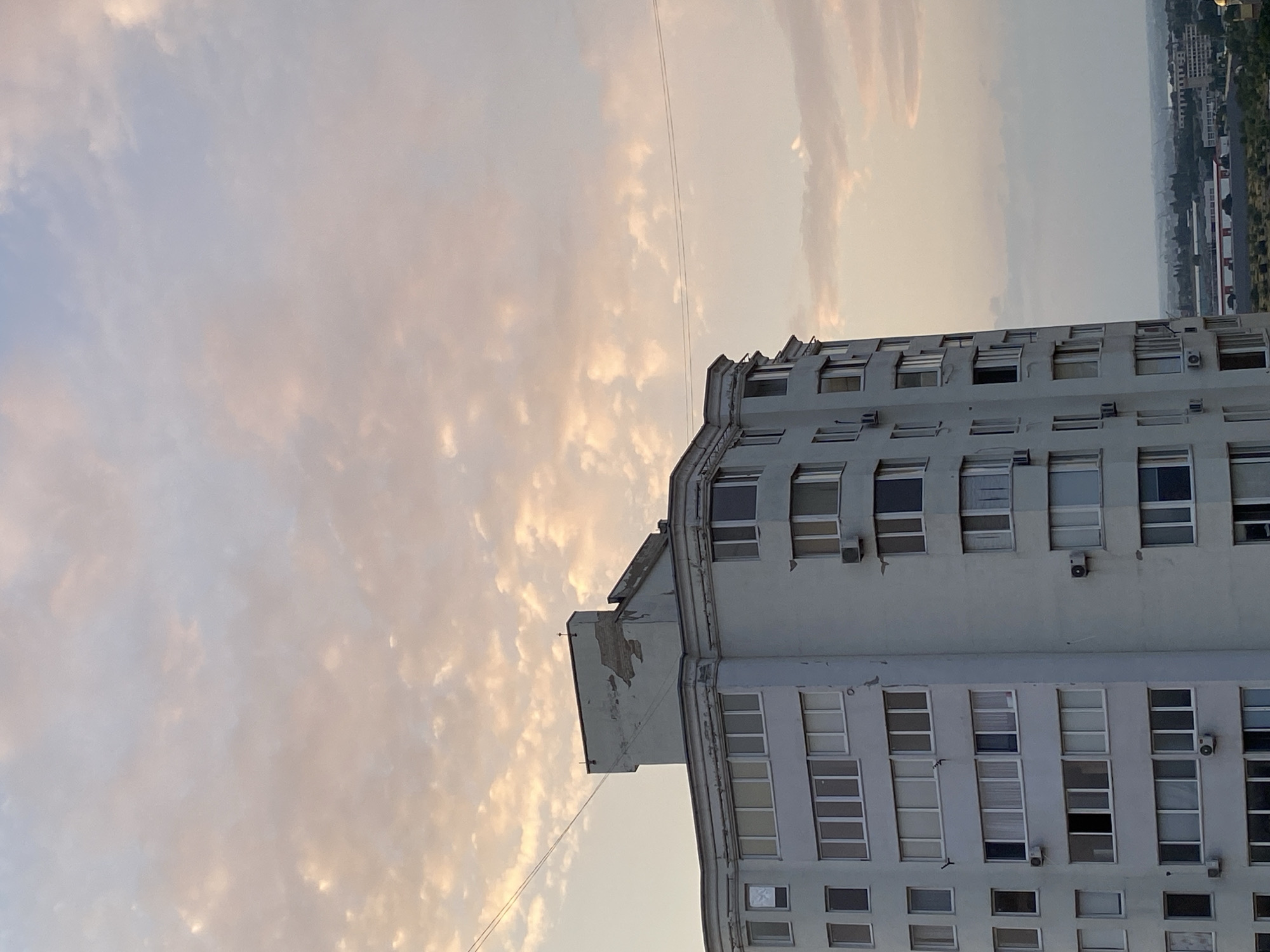 The image shows a multi-story apartment building in Chornomorsk, Ukraine, under a sky filled with soft, pastel-colored clouds during what appears to be sunset or sunrise. The building is light-colored and features numerous windows. The architecture suggests a typical Soviet-era residential block, with some signs of wear and tear on the upper facade. Wires are visible stretched across the sky in front of the building. In the distance, a hazy cityscape with industrial elements and other buildings can be seen. No people are visible in the frame.