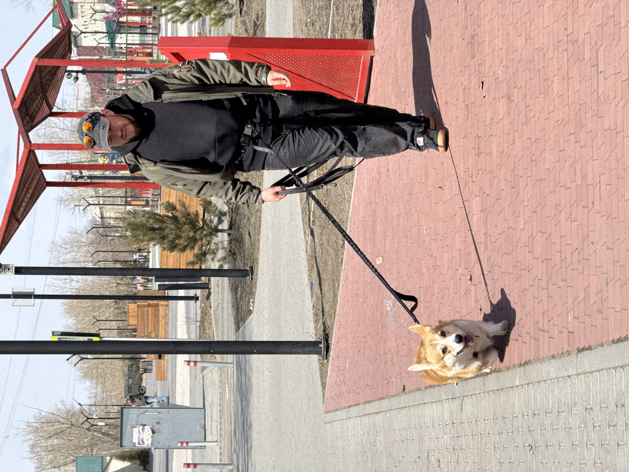 A man walks a corgi on a leash in Borzya, Russia. The man wears a black hoodie, dark pants, a green jacket, sunglasses, and a cap. The dog, a corgi, stands on a paved area. Behind the man is a red structure, and beyond that, trees and buildings are visible. The setting appears to be an urban park or street with trees, walkways, and buildings in the background.
