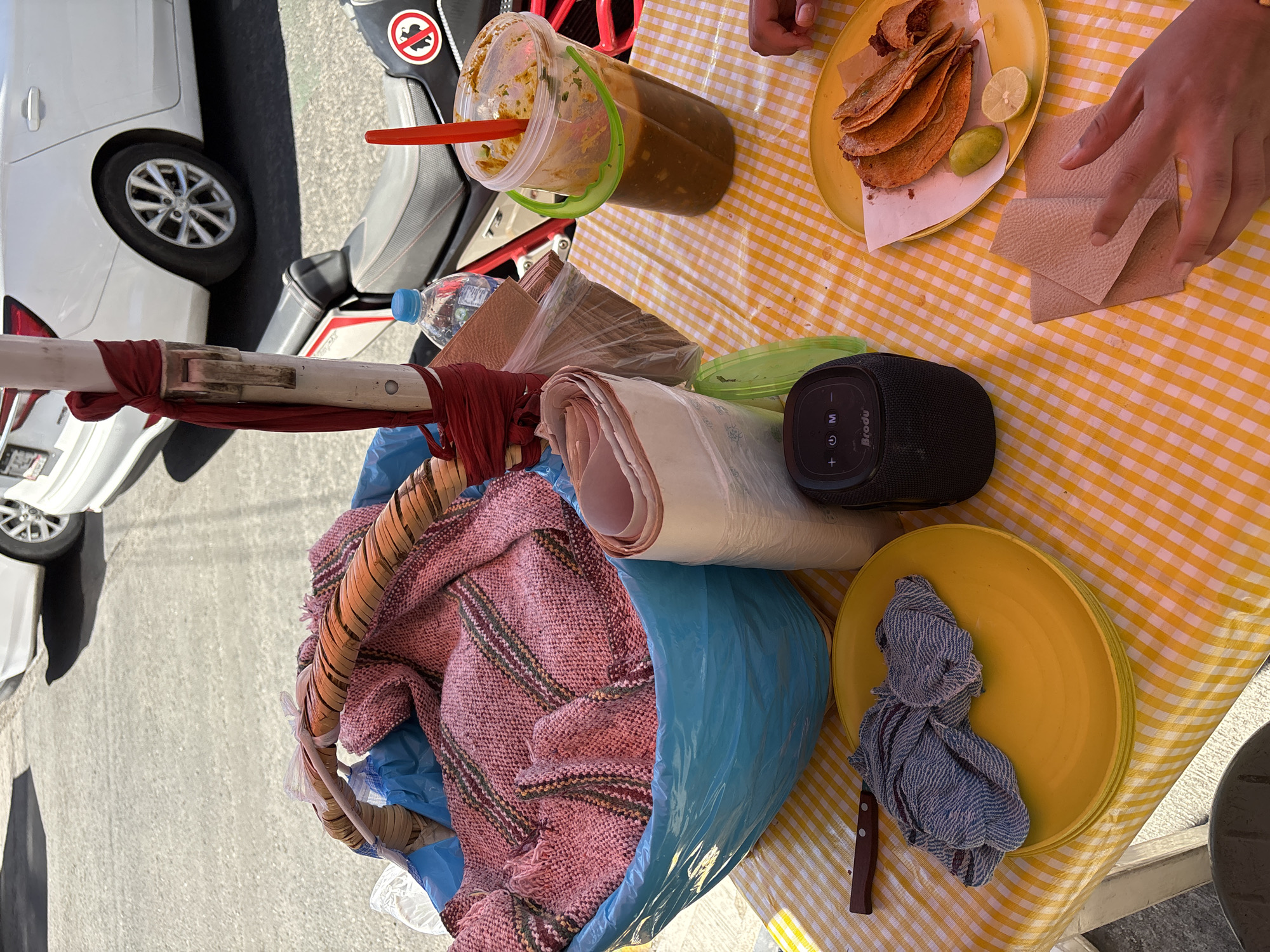 An outdoor scene captures a vibrant street food stand, likely in Puebla, Mexico, during daylight hours. The central focus is a meal featuring several golden-brown tacos, resembling birria tacos, served on a yellow plate with lime wedges. A person's hand is visible to the right, either taking or arranging paper napkins.
Beside the plate, a large plastic container holds a dark reddish-brown liquid, likely consommé or a large batch of salsa, with a red ladle or spoon. A bottled water is also present. The table is covered with a bright yellow and white checkered tablecloth. Other items on the table include a black portable speaker branded "Broodu," a stack of yellow plates, a knife with a wooden handle, and a blue and white striped cloth.
To the left, a white pole, possibly part of the stand's structure, is wrapped with red fabric. Below the table, a blue plastic bag contains what appears to be a pink/red knitted blanket inside a wicker basket, along with rolls of paper or plastic wrap, suggesting supplies or personal items. In the background, a white car and a gray scooter or motorcycle are parked on asphalt. The scooter features a circular "no dogs" symbol, which is a common icon in Mexico. The bright lighting indicates a sunny day.