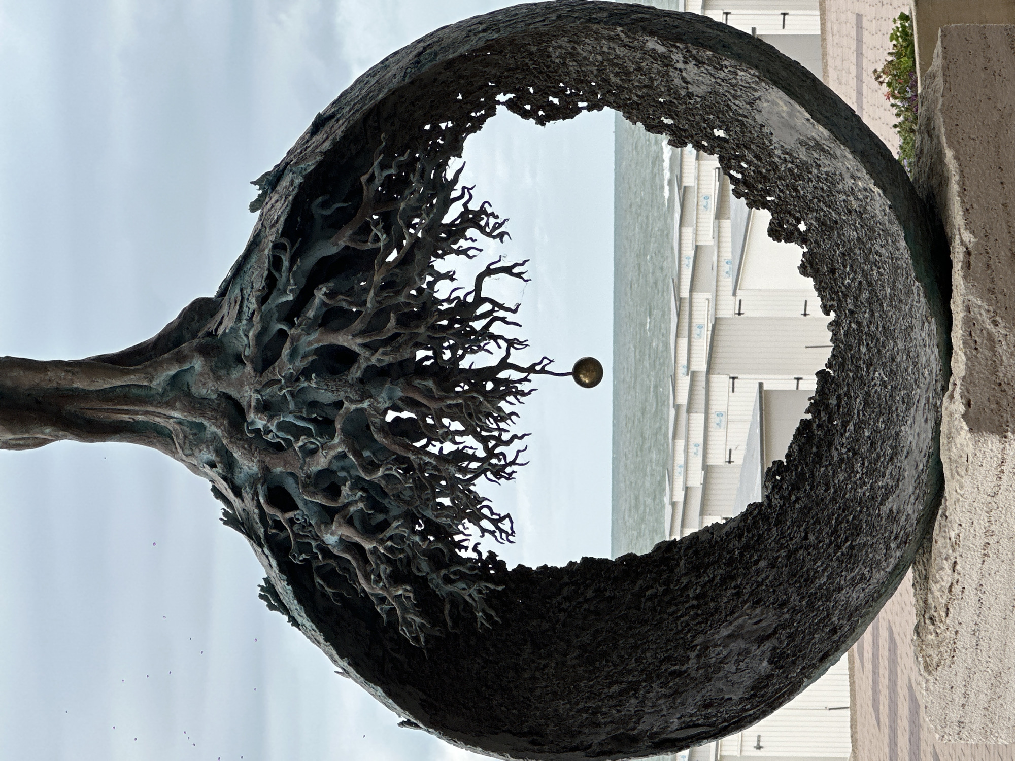 The image displays a bronze sculpture of a tree with exposed roots, forming a circular frame. A golden sphere hangs from one of the roots in the center of the frame. In the background, the sea stretches out, with a row of white beach huts lining the shore. The sky is overcast. The sculpture is situated in front of the beach huts, suggesting a coastal location. This is the "Tree of Life" sculpture by Willibrordus S. S. van der Steen, located on the Zeedijk in Knokke-Heist, Belgium. There are no people or animals visible in the image. The scene is static and captures the sculpture and its surroundings from a frontal perspective.