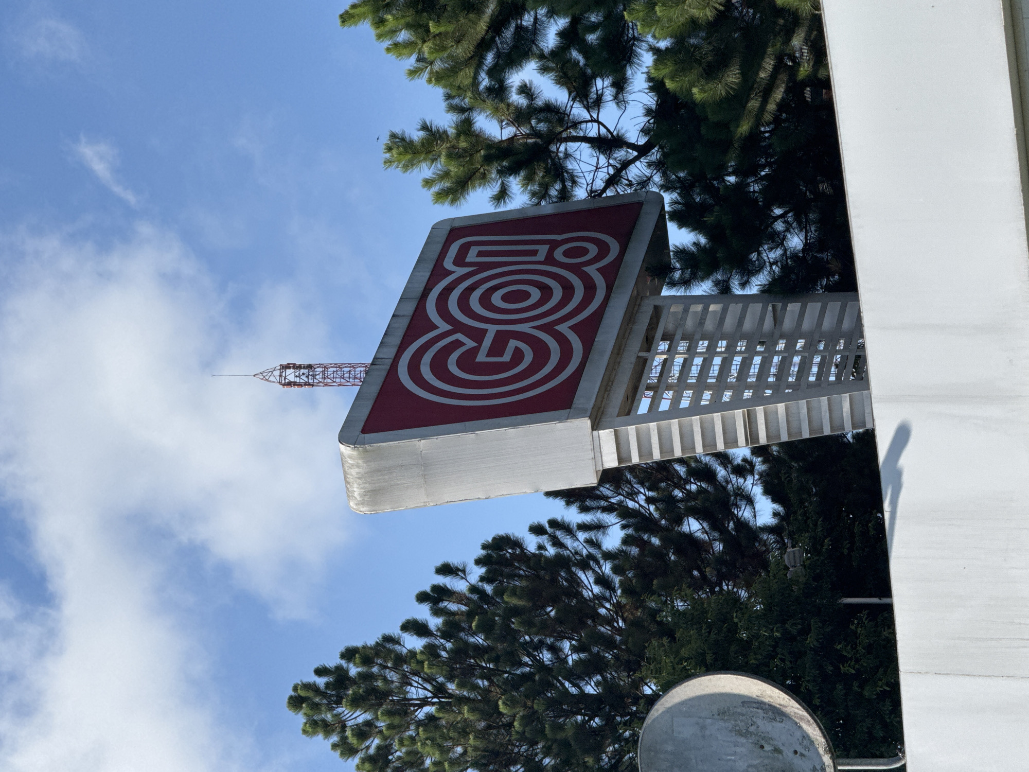 The image shows a sign for "GO!" in Da Lat, Vietnam. The sign is red with white concentric circles forming the letters. It's mounted on a white metal structure. A communication tower is visible in the background. The sky is blue with some clouds. Trees surround the sign. A portion of a building and a traffic mirror are in the foreground.