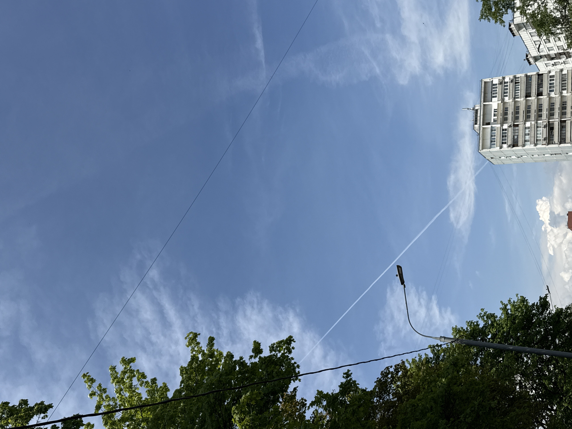 The image shows a view looking up into a blue sky in Moscow, Russia. Thin clouds streak across the sky, and two distinct contrails intersect high above. A tall, white residential building with multiple rows of balconies is visible in the upper right corner. In the foreground, lush green treetops fill the bottom of the frame, partially obscuring power lines and a black street lamp.