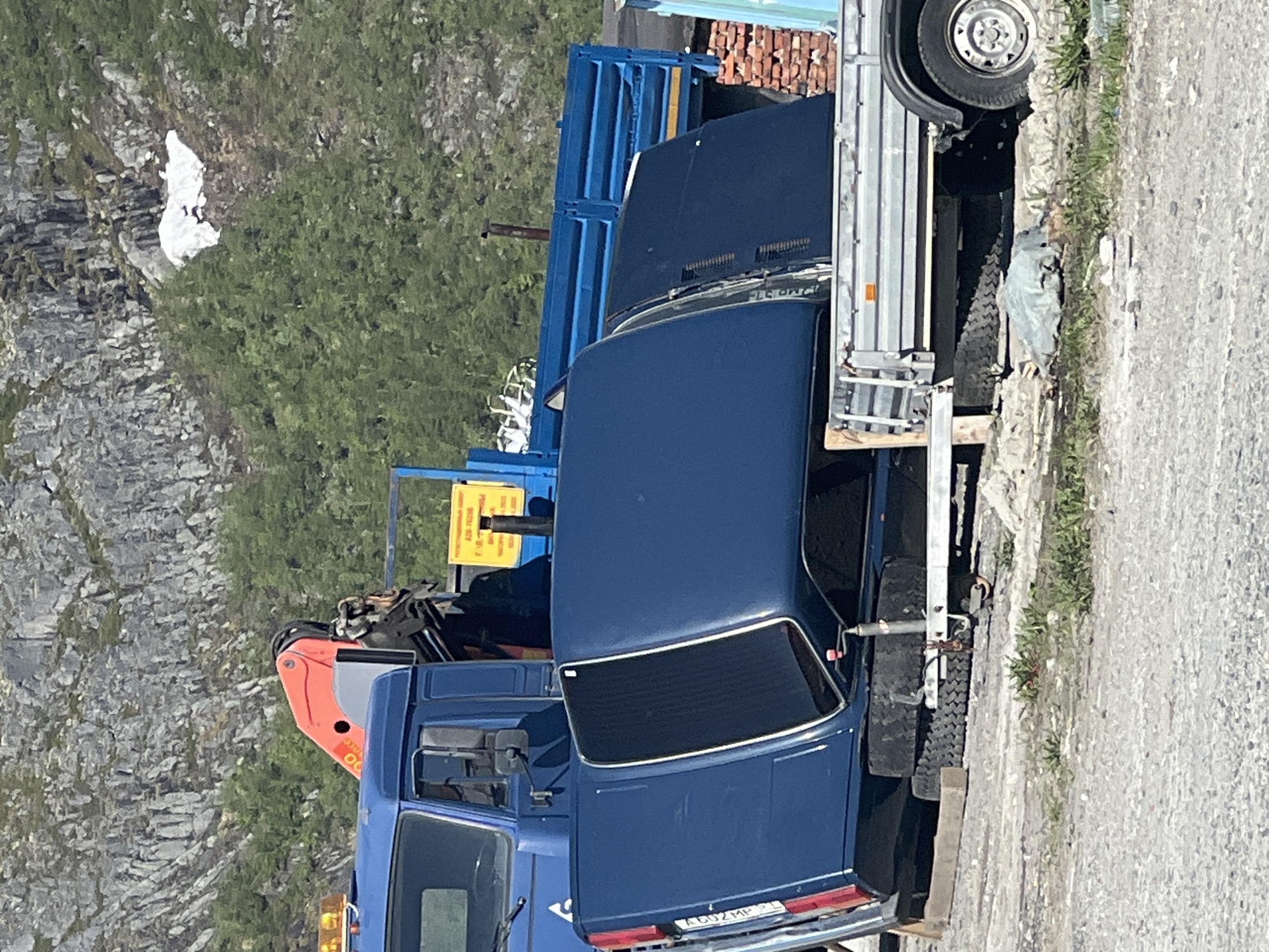 In Kirovsk, Russia, an outdoor scene depicts a blue truck with a crane attachment parked on a gravel surface. The truck's bed is covered by a blue panel, and another blue panel is resting on top of it. Behind the truck, a blue trailer is visible, loaded with various items. In the background, a rocky mountainside with patches of snow and green trees rises under a clear sky.