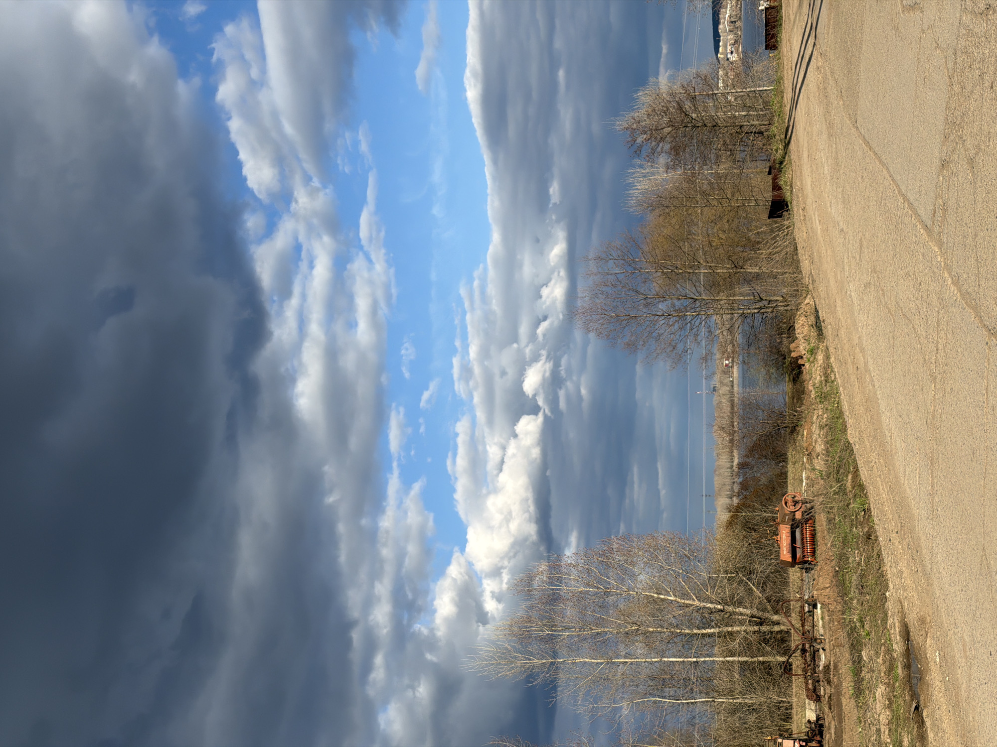 This image presents a dramatic outdoor landscape, likely in Noviy, Russia, characterized by a striking sky and a semi-rural setting.
The dominant feature is the sky, which exhibits a sharp dichotomy: the right half of the frame features patches of clear blue sky interspersed with bright white, fluffy clouds, while the left half is consumed by a large mass of dark, heavy storm clouds, suggesting an approaching or receding weather front.
Below the sky, the landscape consists of a light-colored, unpaved or gravel road traversing diagonally across the right side of the frame, leading towards the distance. Adjacent to the road, the ground is covered with dry grass and bare earth. Several deciduous trees, stripped of their leaves, stand prominently in the middle ground, indicating late winter or early spring. Their bare branches form intricate patterns against the sky and water.
Further into the middle ground on the left, an old, rusty piece of machinery, possibly an agricultural implement like a hay rake, is partially visible, adding a rustic element to the scene. Beyond the trees and machinery, a calm body of water, likely a river or lake, stretches across the horizon. On the far bank of this water, distant trees and indistinct structures can be faintly seen.
No people are present, and no particular activity or event is occurring; the image is a static portrayal of a natural environment. The dramatic contrast in the sky's appearance, from clear to stormy, is a notable detail, creating a sense of dynamic weather. The leafless trees and rustic machinery contribute to an atmosphere of quiet rural life. There is no visible text in the image.