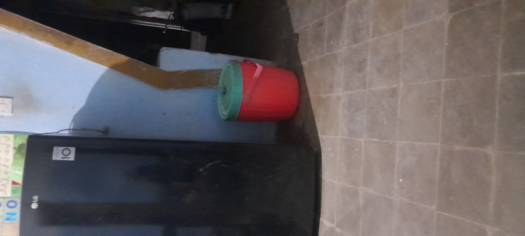 A black LG refrigerator stands to the left of the frame. Above the refrigerator, a sign is partially visible with the letters "NO" in blue and white. To the right of the refrigerator, a red bucket with a green lid and handle is placed on a tiled floor. The walls in the background are painted light blue and feature a diagonal yellow stripe. The scene appears to be indoors, possibly in a residential or commercial space. There are no people or animals visible in the image. The floor is covered in square beige tiles. The image is taken from a medium distance, showing a significant portion of the refrigerator and the bucket.