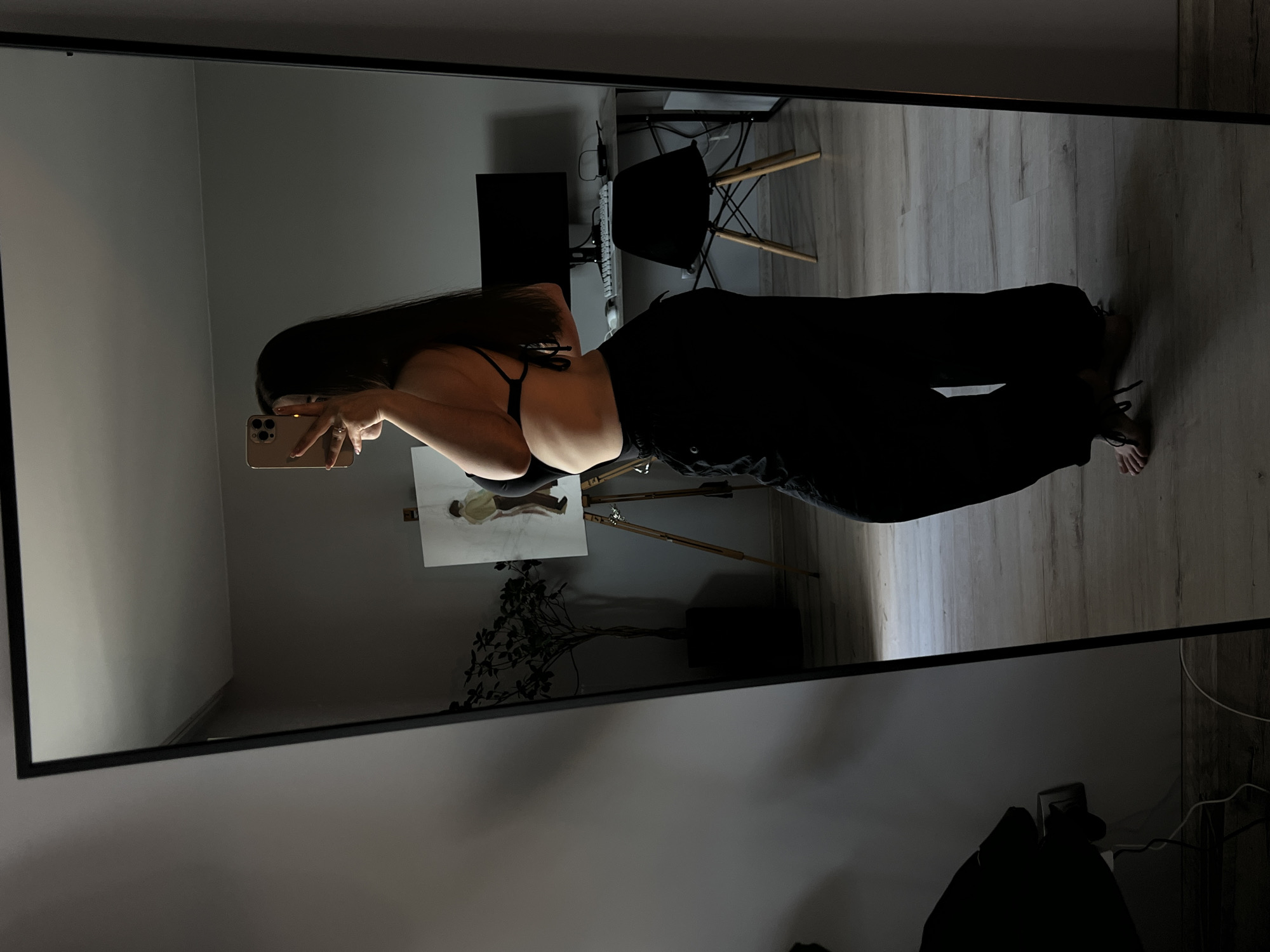 The media file shows a full-length mirror reflecting a young woman in a black bikini top and black wide-leg pants. She is holding a phone in her right hand, seemingly taking a selfie. Her long, dark hair is draped over her shoulders. The reflection also reveals a studio-like setting with a desk, a computer monitor, a keyboard, and a black chair. An easel with a painting and a potted plant are visible in the background, suggesting an artistic or creative space. The floor is made of light-colored wood planks. The lighting appears to be artificial, casting shadows and creating a moody atmosphere. The overall mood seems casual and relaxed. The location context of Ternopil, Ukraine is provided. There is no visible text in the image.
