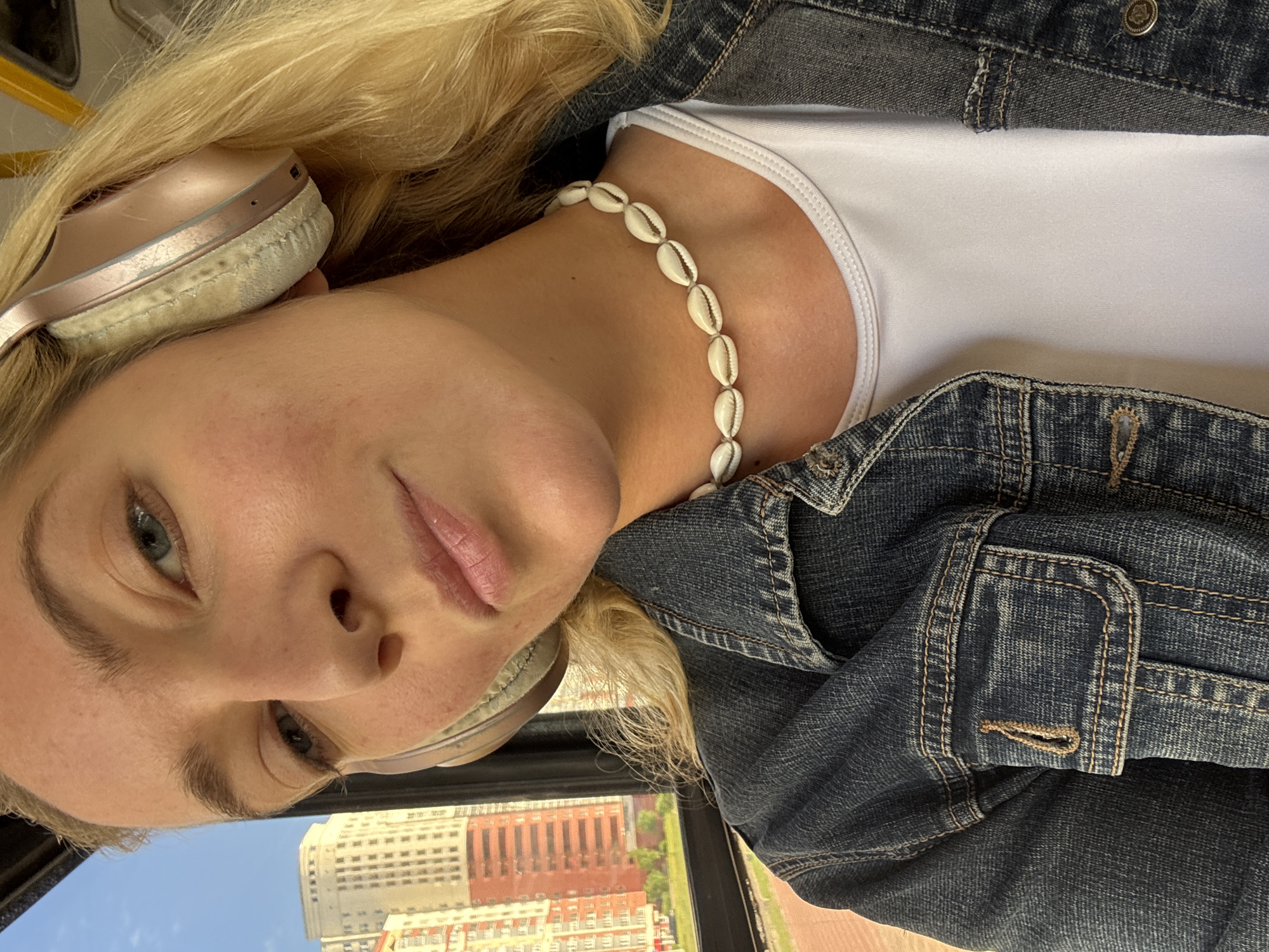 A young blonde woman is captured in a selfie, sitting in what appears to be a bus or train, with a city skyline visible through the window behind her. She is wearing rose gold headphones over her ears and a white shell necklace. Her clothing consists of a white t-shirt and a dark denim jacket. The background shows a building with distinctive orange-red horizontal bands, characteristic of Soviet-era architecture, which is common in Minsk, Belarus. The lighting suggests it is daytime.