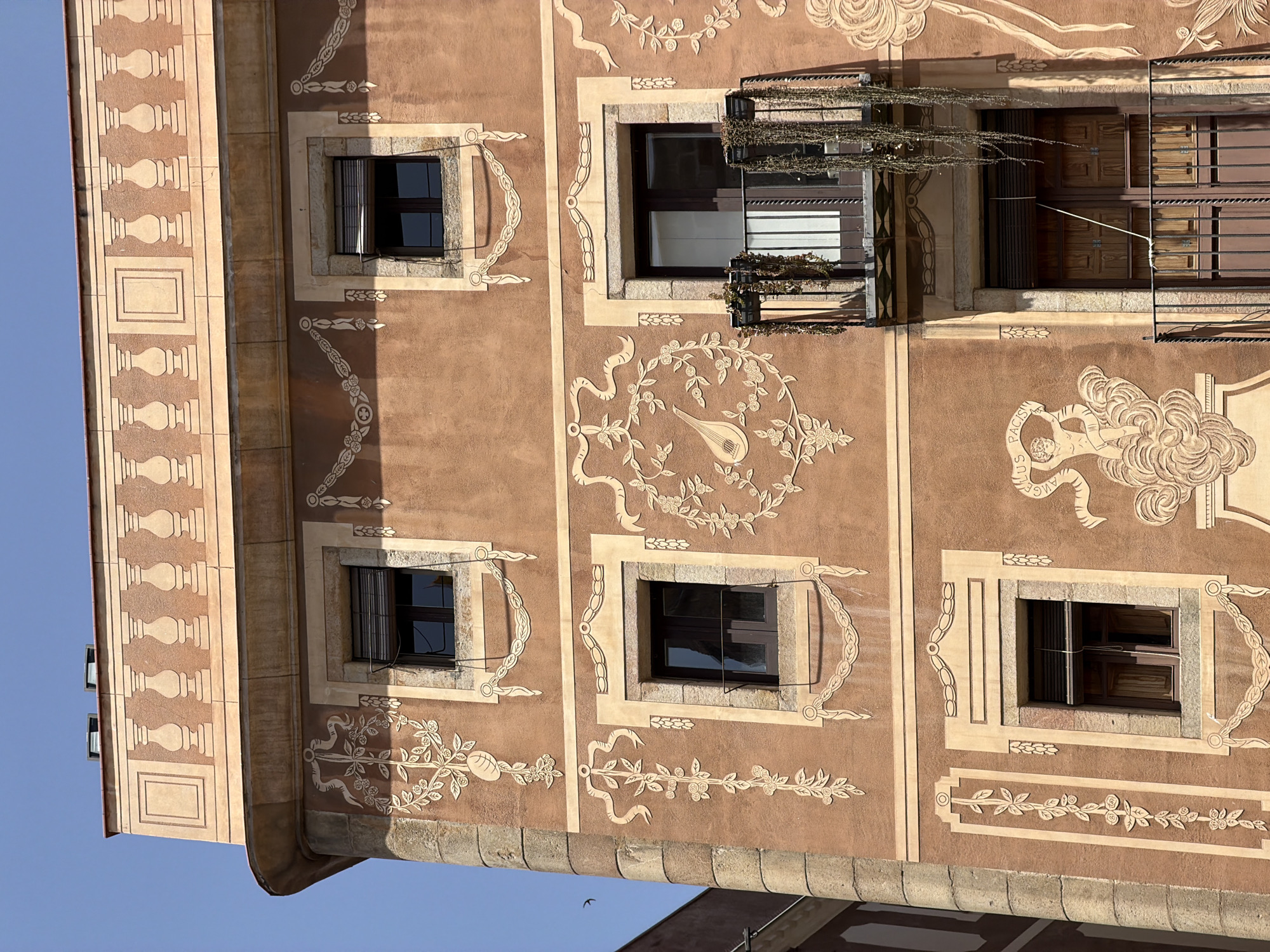 The image captures a detailed view of the facade of a building in Barcelona, Spain, likely a residential or commercial structure, characterized by its ornate architectural details. The building's exterior is rendered in a warm, terracotta-like hue, adorned with elaborate, light-colored decorative plasterwork. This ornamentation includes intricate floral motifs, garlands, ribbons, and classical elements such as balustrades and scrolled designs.
There are several windows visible, some with dark frames and shutters, and one prominent balcony with black metal railings and hanging plants. The balcony features a wrought-iron structure and appears to be attached to a doorway. Above the windows and extending across the top of the facade is a decorative frieze, mimicking a classical balustrade with alternating pillar and panel designs. The sky above is clear and blue, indicating fair weather.
The architectural style suggests a building from the late 19th or early 20th century, potentially influenced by Art Nouveau or a revivalist style common in Barcelona. The level of detail in the plasterwork is a key feature, giving the building a rich and historic appearance. The composition is a close-up, focusing on the upper floors of the building, highlighting the decorative elements and the architectural features. The image is taken from a slightly upward angle, emphasizing the scale and height of the structure. A small bird is visible flying in the sky to the left of the building.