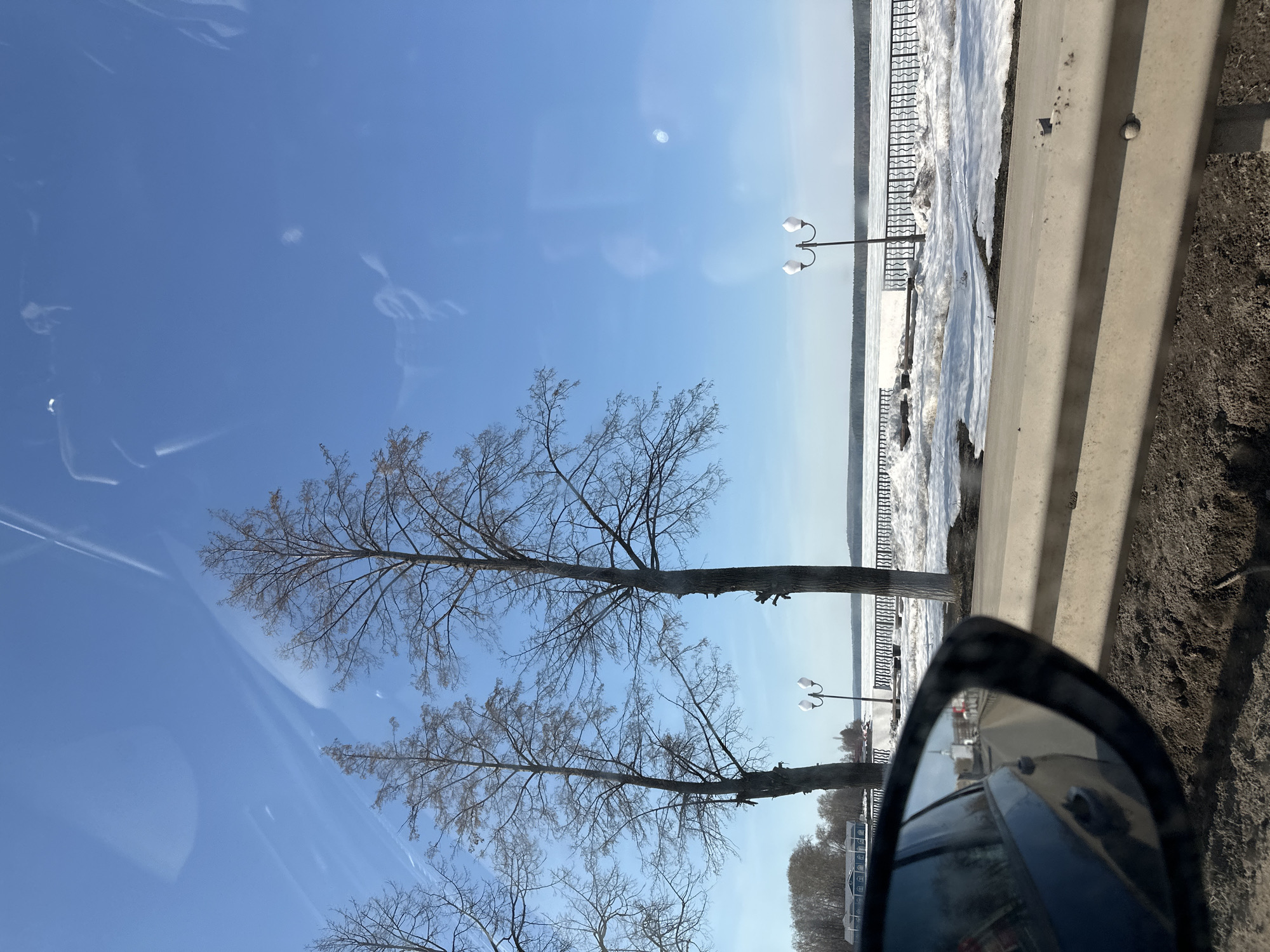 The image, taken from inside a car, shows a roadside view in Votkinsk, Russia. Through the windshield and side mirror, a clear blue sky is visible above bare trees. A light pole stands alongside a concrete barrier near a partially frozen river or lake. The side mirror reflects traffic and urban elements, indicating the car is in a populated area. Snow and ice accumulate near the barrier.