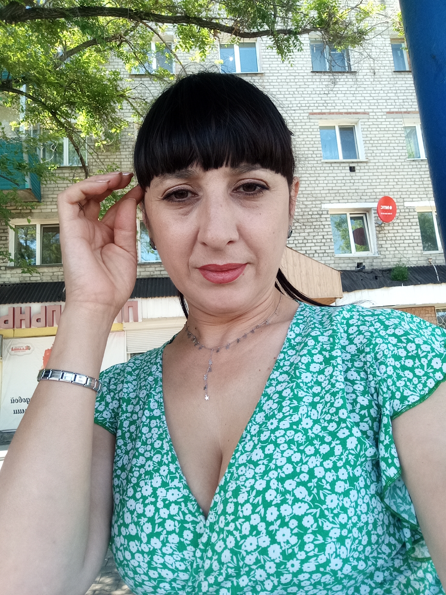 A middle-aged woman with dark hair and bangs, wearing a green floral dress and a silver bracelet, poses for a selfie outdoors. She is looking at the camera with a slight smile. Her right hand is raised to her head, touching her hair. Behind her, a light-colored brick building with several windows is visible, along with green trees and a red satellite dish. The location is identified as Belogorsk, Russia.