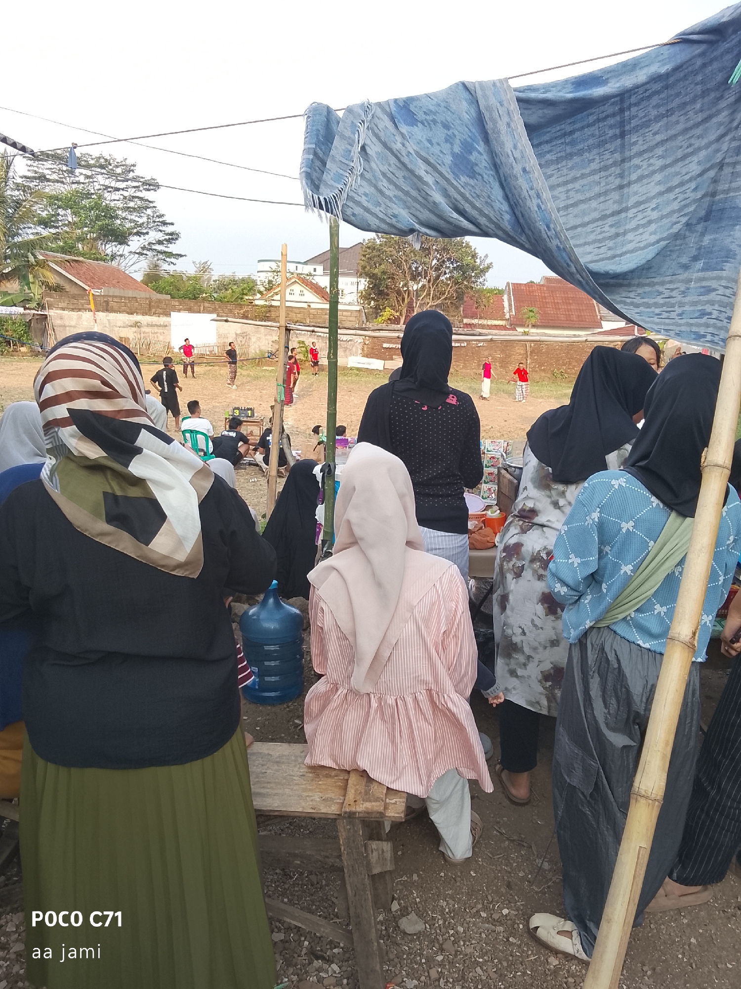 The image captures an outdoor gathering where several individuals, predominantly women wearing headscarves, are observed. In the foreground, a woman is seen from behind, her head adorned with a patterned headscarf, and she is wearing a black top and a long olive-green skirt. To her right, another woman in a light pink, striped, peplum-style top and light-colored pants is seated on a wooden bench. Further to the right, several other women are visible, one in a grey and white patterned dress and another in a blue patterned top and dark pants. A large blue water cooler jug is situated near the woman in the foreground. In the background, a group of men and boys are playing what appears to be a game, possibly volleyball, in a dirt field. The scene is partially covered by a large blue tarp supported by bamboo poles, suggesting a temporary shelter or vendor stall. The environment appears to be a somewhat rural or semi-urban area, with buildings and trees visible in the distance. The sky is overcast. The provided context indicates the location is in the city of Tasikmalaya, Indonesia.
