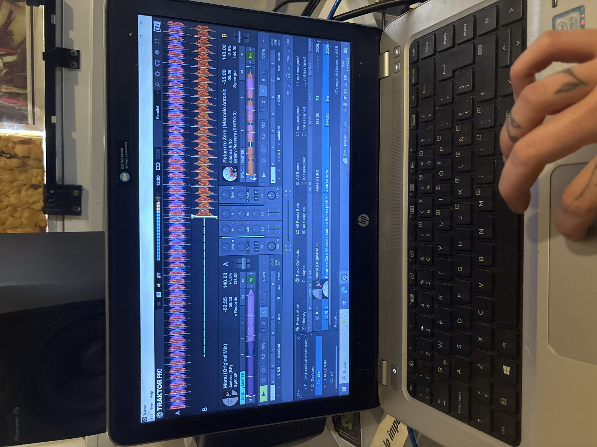 A laptop is on a table. The screen displays Traktor Pro software, a DJ application. The software shows waveform visualizations of audio tracks, with song titles "Moral (Original Mix)" by Anders (BR) and "Return to Zero (Marcelo Antonic Remix)." The laptop keyboard is visible, and a hand with tattoos is visible on the right edge, using the trackpad. The location is Buenos Aires, Argentina, indicated by the weather information in the bottom right corner, which reads "21°C Mayorm. nubla..."