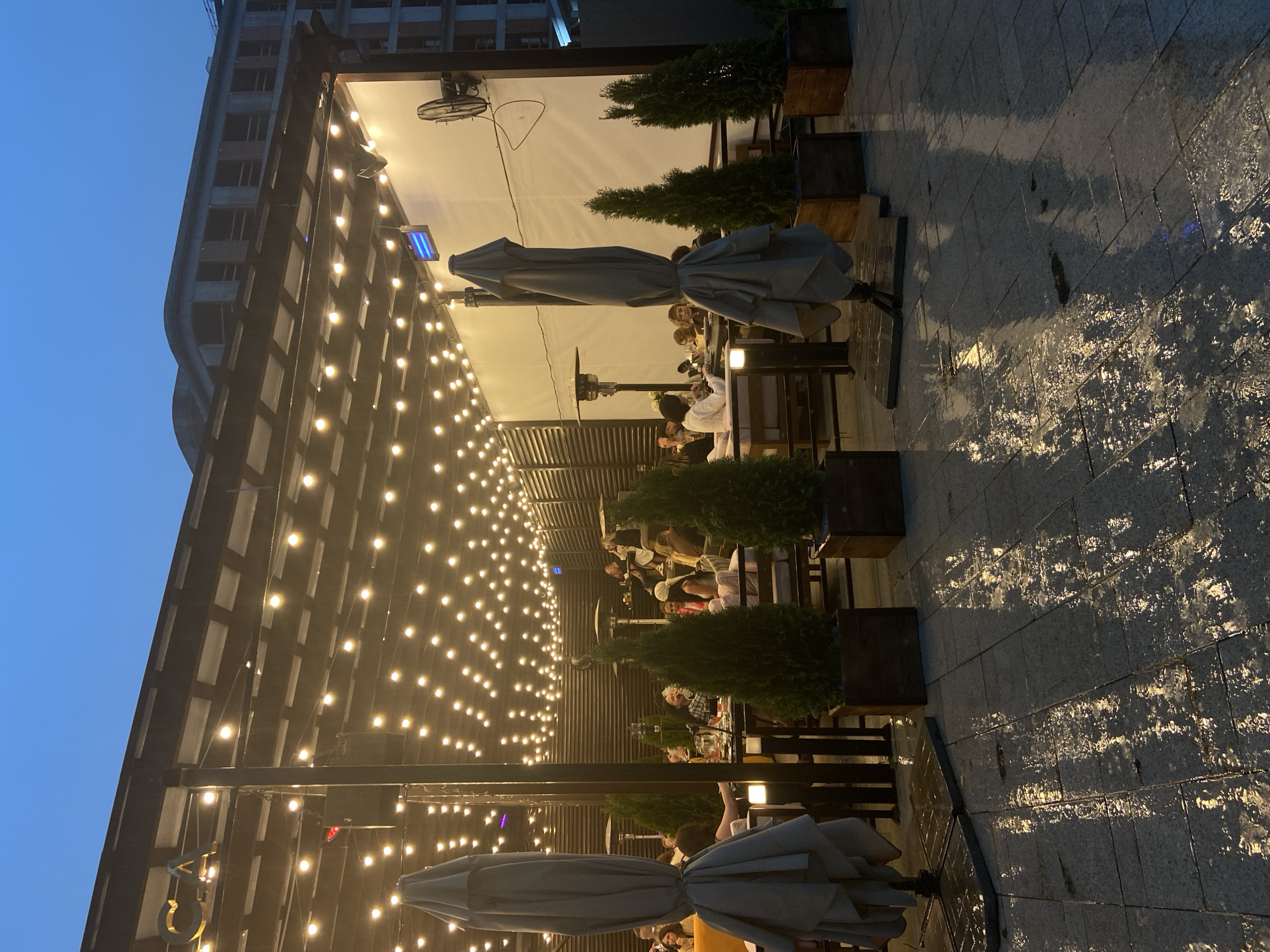 This image depicts the outdoor dining area of CAVA Lounge Cafe, located at Lenin Street 67/10 in Tyumen, Russia, during the evening or night. The sky above is a dark blue, indicating twilight or nighttime. The ground, paved with grey rectangular tiles, is wet and highly reflective, suggesting recent rainfall or damp conditions.
The scene focuses on a covered patio structure supported by dark columns and beams, featuring an overhead canopy strung with numerous warm yellow incandescent lights. These lights cast reflections on the wet pavement. Along one side of the patio is a dark wall with horizontal slats, and on the opposite side, a plain white wall. Attached to the white wall are at least two blue-glowing electric heaters and one black wall-mounted oscillating fan.
Within the patio area, approximately 15 to 20 individuals, predominantly adults, are seated at various tables, engaging in conversation and dining. Their attire appears casual to semi-casual. Several tall, folded grey patio umbrellas are positioned throughout the space, along with multiple freestanding patio heaters. Potted evergreen plants in square wooden planters are strategically placed as decor or dividers between seating arrangements. In the background, against the dark sky, a large, multi-story building with exposed concrete and numerous rectangular window openings is visible, appearing utilitarian or under construction.
