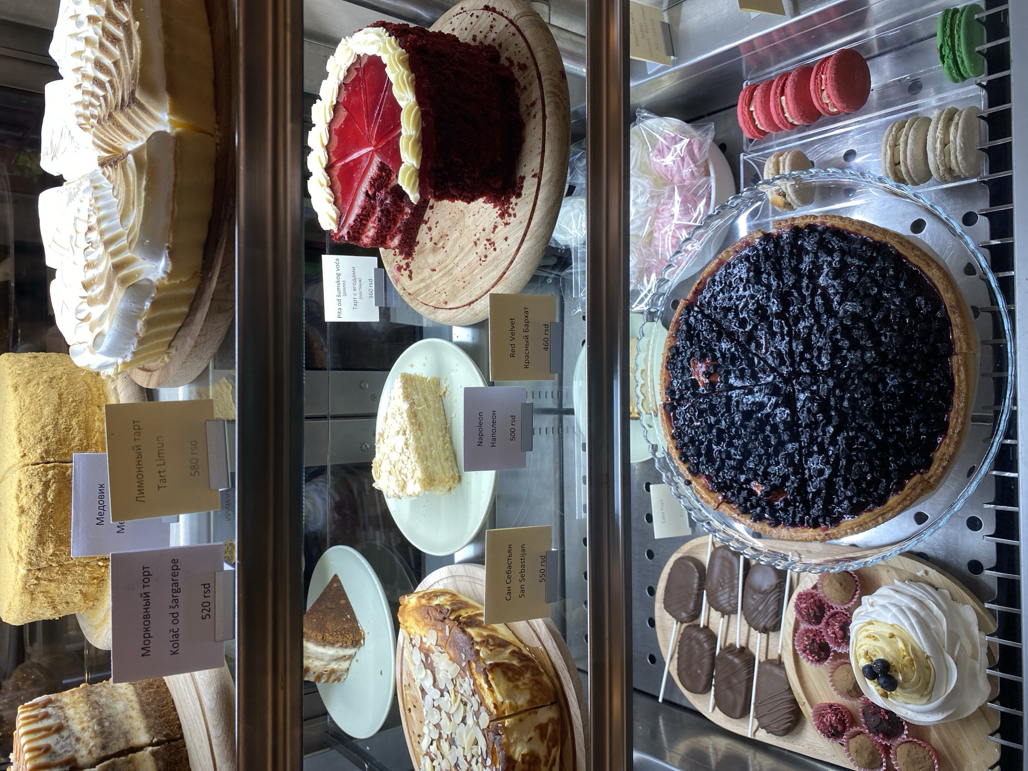 The image captures a display case filled with various cakes and pastries, likely inside a bakery or café in Novi Sad, Serbia.
On the left, there's a multi-layered cake topped with a creamy meringue-like frosting. Below it, a square-cut honey cake ("Medovik") is placed. A carrot cake ("Morkovni tort") is also visible.
In the center section, there is a slice of Red Velvet cake, a piece of Napoleon cake, and San Sebastian cake.
The right section showcases a blueberry tart. Below it is a selection of desserts like cake pops and meringue.
The cakes are all labeled with tags in Serbian, displaying their names and prices in Serbian dinars (rsd). Macarons are lined up at the top of the display case. The scene suggests a bakery or cafe offering a diverse range of desserts.