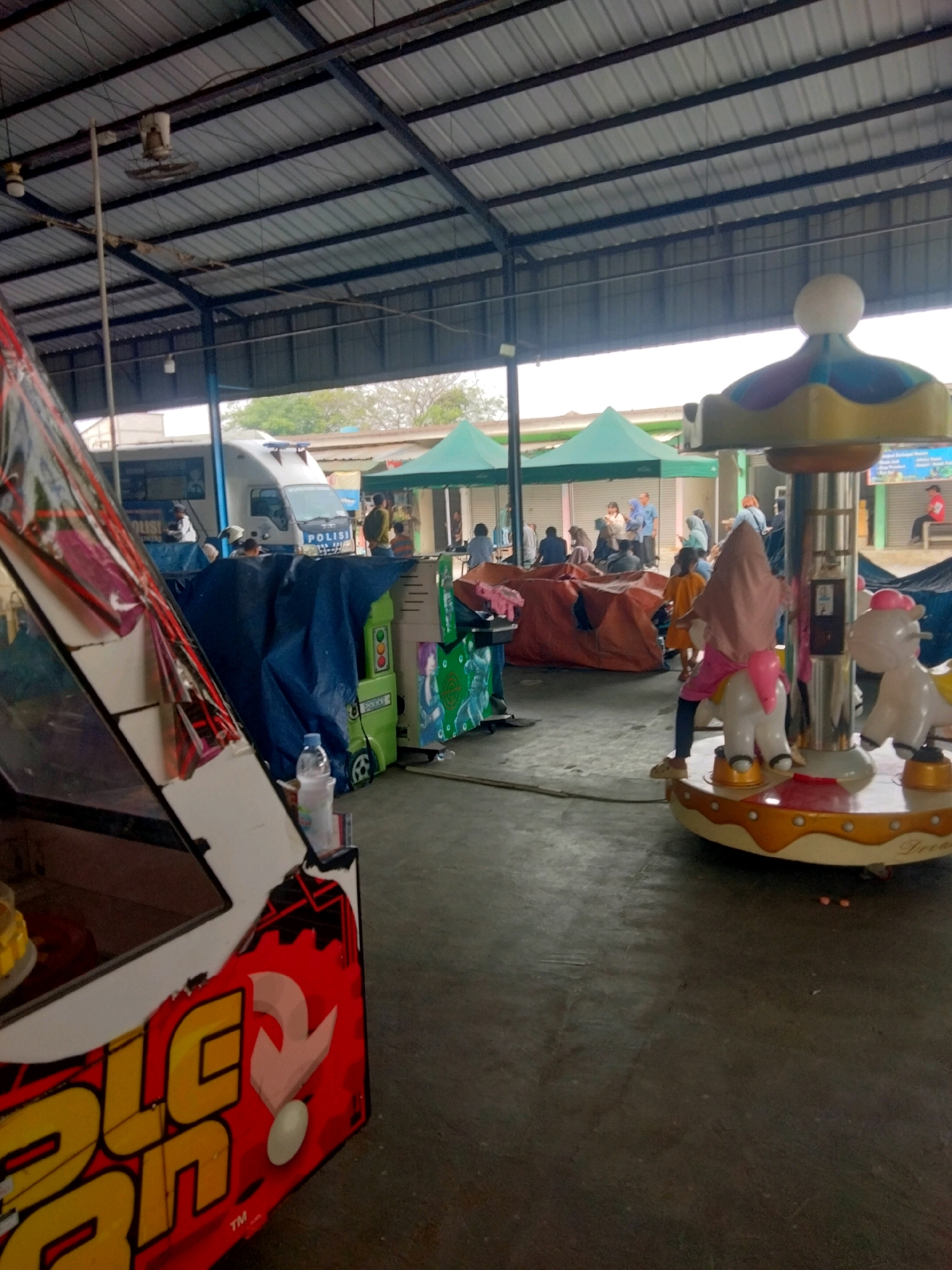 The image depicts an outdoor, covered area, possibly a marketplace or an amusement park, in Sukaasih, Indonesia. In the foreground on the left, a large, red and yellow arcade game machine with a cracked screen is visible. To the right of the arcade machine, a small green children's ride is covered with a dark blue tarp. Further to the right, a carousel with white horse figures is in operation, with a young child wearing a pink hijab and a pink shirt riding one of the horses.
In the background, several people are milling about. A white police van with "POLISI" written on it is parked behind a blue tarp. Several green tents are set up in the mid-ground, and beyond them, buildings and trees are visible. The roof of the structure is made of corrugated metal supported by black metal beams. The ground is paved with concrete. The overall scene suggests a busy, informal gathering or recreational area.