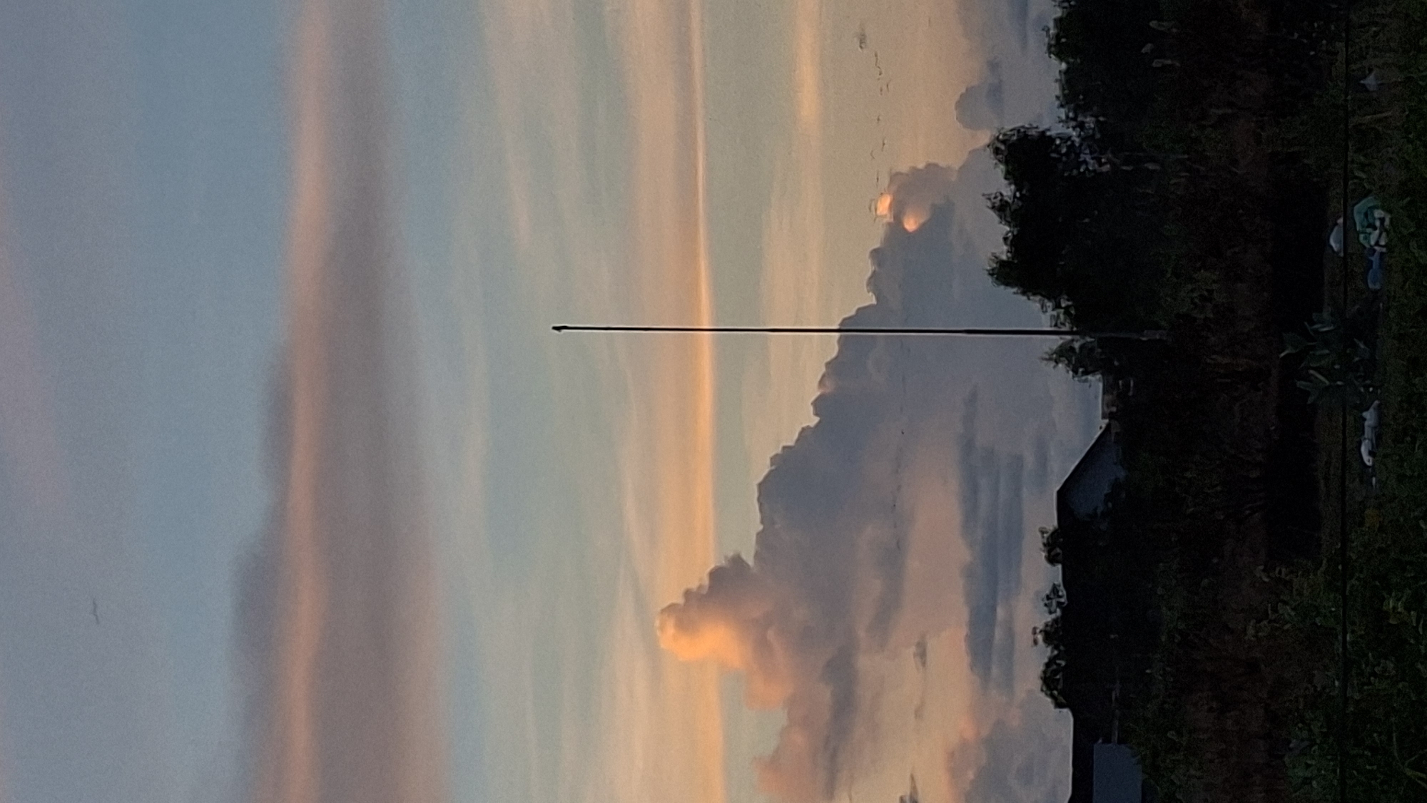 This image captures a twilight scene in Lhokseumawe, Indonesia, dominated by a dramatic sky. The sky displays a gradient of colors, transitioning from soft blues and pinks near the horizon to deeper blues and grays higher up. Wispy clouds are scattered across the sky, with some larger, more defined cumulus clouds in the middle ground, illuminated by the setting sun, giving them a warm, orangey-pink glow.
In the foreground, silhouetted against the vibrant sky, is a dense line of trees and foliage. A thin, dark pole, possibly an antenna or a utility pole, stands vertically in the center-right of the frame, extending from the ground into the sky. A few indistinct buildings are visible through the trees, suggesting a residential or semi-urban area. The overall impression is one of a quiet evening unfolding in Lhokseumawe.