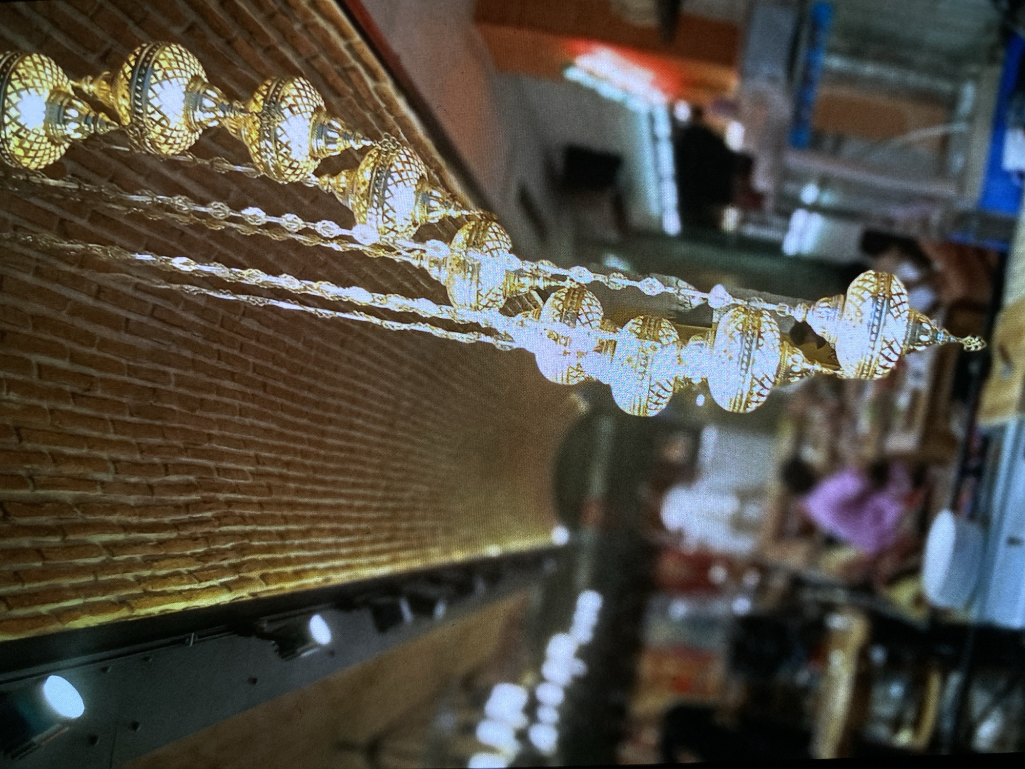 This media file depicts an indoor scene in Bankya, Bulgaria, dominated by a decorative string of ornate golden or brass-colored lights. These intricate lamps or lanterns, featuring detailed latticework, are illuminated and hang diagonally across the frame, connected by delicate chains, casting a warm, inviting glow.
To the left, a brick wall forms a textured background, subtly lit from below by a row of visible spotlights or track lighting mounted on a dark rail. This upward illumination highlights the brickwork and complements the warm tones of the hanging lights.
The right side of the image features a blurred background, suggesting a bustling indoor environment. Indistinct figures of people are visible, including one person wearing purple clothing. The blurry shapes indicate various objects, possibly furniture, merchandise, or other elements typical of a commercial or hospitality setting like a restaurant, cafe, or shop.
The scene conveys a cozy and potentially elegant or festive atmosphere, likely during the evening or night, given the prominent use of artificial lighting. No discernible text is visible in the image.