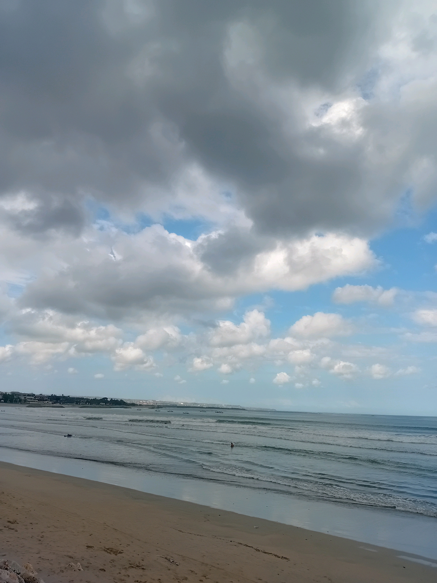 The image captures a wide view of Kuta Beach in Kuta, Indonesia. The sky is filled with dramatic, gray clouds mixed with patches of bright blue sky, indicating a possibly overcast or transitioning weather day. The ocean stretches out to the horizon, with gentle waves rolling onto the sandy shore. Several small boats are visible in the distance along the coastline, where buildings and vegetation form the backdrop of the city. On the beach, the sand is a light brown color with some scattered debris. There are a few people visible in the water, one appearing to be swimming or wading, and another on a small watercraft, suggesting recreational activities are taking place. The overall scene depicts a dynamic coastal environment with natural elements like the sky and sea dominating the frame.