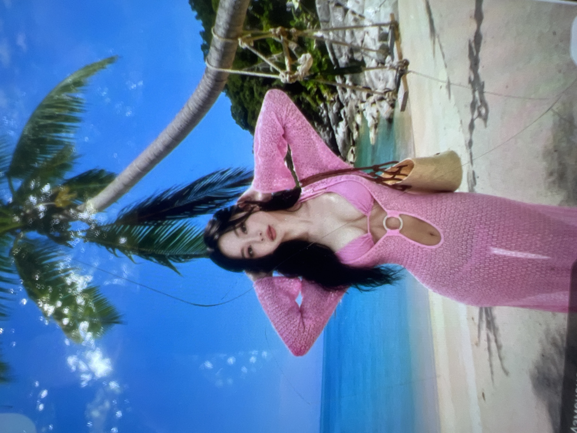 A woman with long, dark hair is posing on a beach. She is wearing a pink bikini and a pink knitted dress with cutouts. She has her hands on her head and is looking at the camera. The background is a tropical beach scene with a palm tree, a hammock, and the ocean. The location is not Kara-Dıykan, Kyrgyzstan; it is a beach.