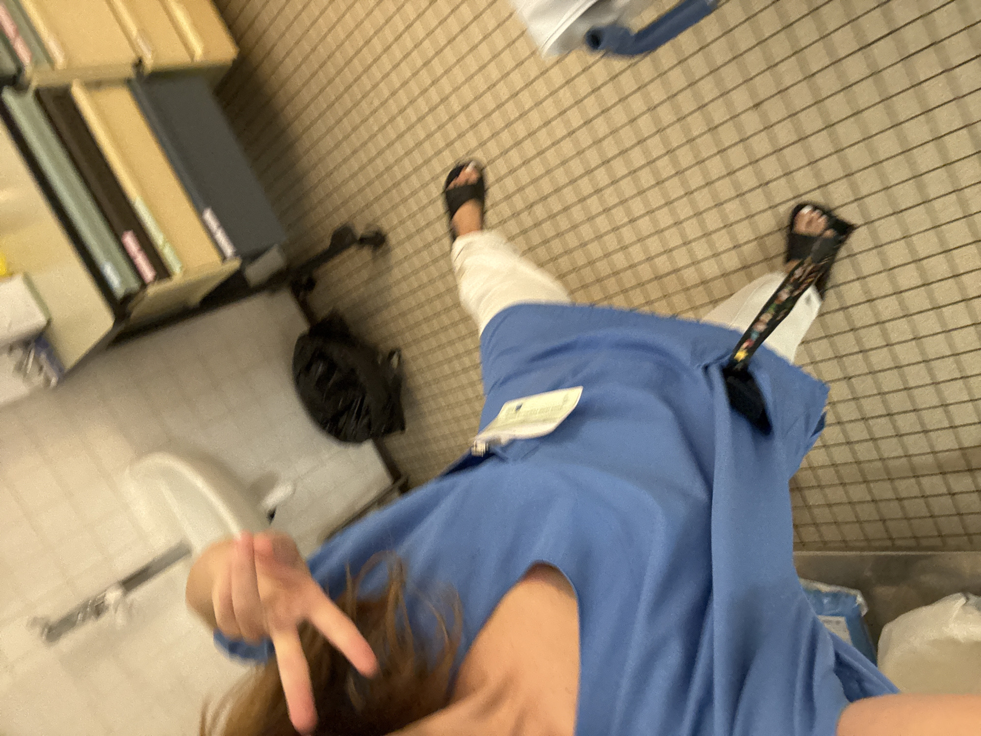 The image shows a person in blue scrubs, possibly a healthcare worker, in what appears to be a utility room or storage area. The person is giving a peace sign with their left hand. In the background, there is a sink and a rolling cart with drawers filled with folders. There is also a black trash bag on the floor. The floor is tiled with a grid pattern. The image does not provide any specific clues about the location in Tourcoing, France.