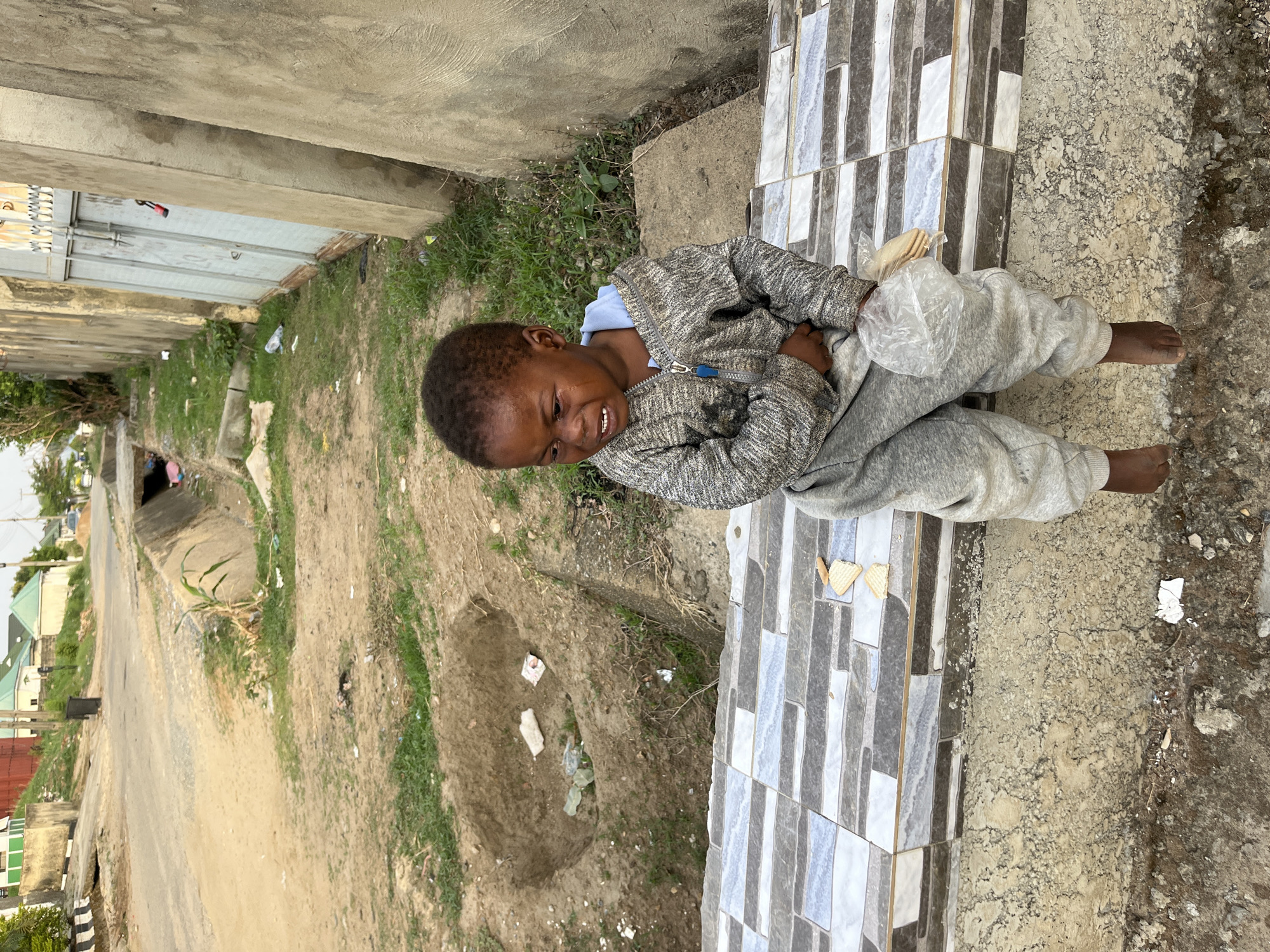 A young, barefoot boy is sitting on a tiled step, looking distressed. He is wearing a gray tracksuit jacket and sweatpants. In his lap, he is holding a plastic bag that appears to contain some food items. The tiled step is gray and white. The surroundings appear to be a street scene in Abuja, Nigeria. The ground is dirt and grass with some litter. In the background, there is a street and several houses. The location seems to be an urban or peri-urban area in Abuja.