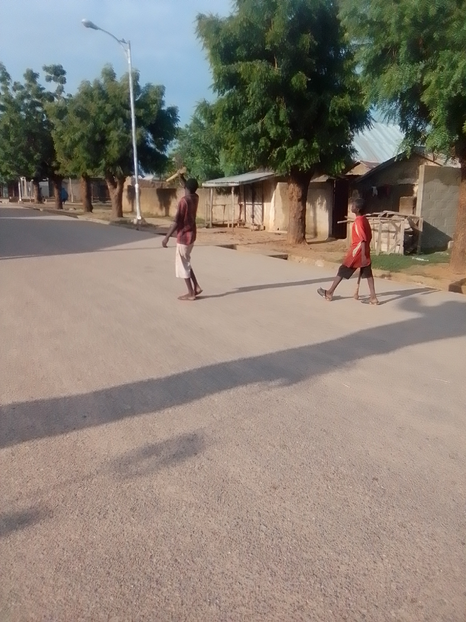 In Liji Hill, Nigeria, two young individuals walk on a paved road adjacent to trees and buildings. One person, wearing a maroon shirt and light-colored shorts, walks towards the left side of the frame, casting a long shadow. The second person, dressed in a red shirt and dark shorts, is further to the right and also walking. The environment includes a street lamp, several trees with lush green foliage, and some structures that appear to be residential or commercial buildings with corrugated metal roofs. The sky is clear and blue, suggesting daytime.